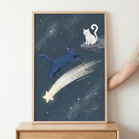 A framed whimsical illustration features two cats in a deep blue celestial setting. A dark blue cat jumps through the air, while a white cat sits calmly on a rocky floating ledge in the upper right. A bright yellow shooting star with a long, glowing tail streaks across the scene. The background is filled with a dense field of stars and cosmic clouds. This art piece is held within a light-colored wooden frame, resting on a wooden surface against a neutral wall.