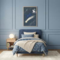 A serene bedroom with a celestial theme features light blue walls accented with classic panel moulding. In the center, a bed with a navy blue upholstered headboard is dressed in a comforter and pillows patterned with white stars. A chunky, cream-colored knit throw is layered over the foot of the bed. 

