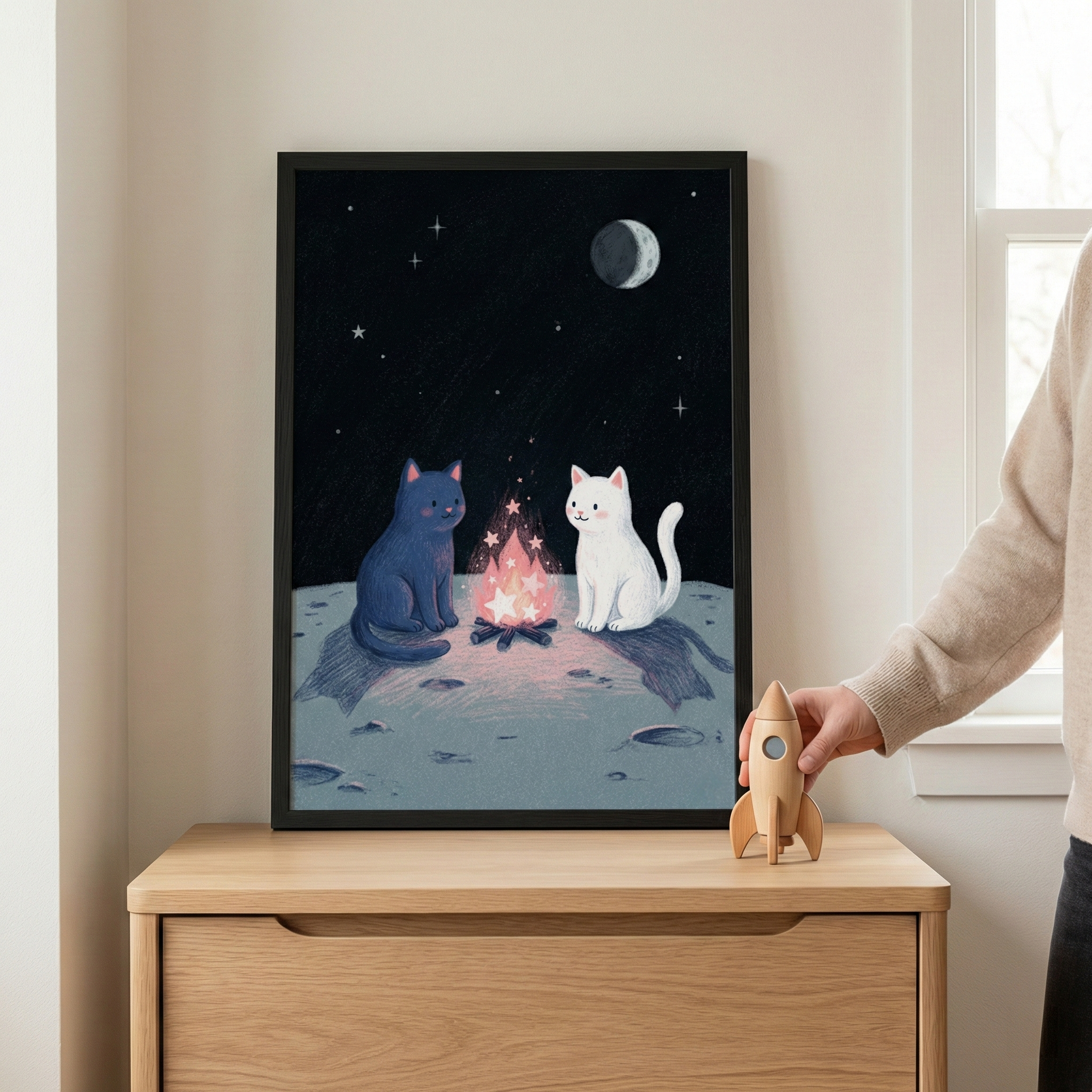 A framed illustration sits on a light-colored wooden cabinet. The artwork features two simple cartoon cats—one navy blue and one white—sitting around a pink campfire on the surface of a moon. The moon is covered in craters, and the dark night sky above is filled with tiny stars and a crescent moon. To the right of the frame, a hand is shown holding a small, unpainted wooden rocket toy. The setting is a bright, minimalist room with a window nearby.