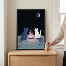 A framed illustration sits on a light-colored wooden cabinet. The artwork features two simple cartoon cats—one navy blue and one white—sitting around a pink campfire on the surface of a moon. The moon is covered in craters, and the dark night sky above is filled with tiny stars and a crescent moon. To the right of the frame, a hand is shown holding a small, unpainted wooden rocket toy. The setting is a bright, minimalist room with a window nearby.
