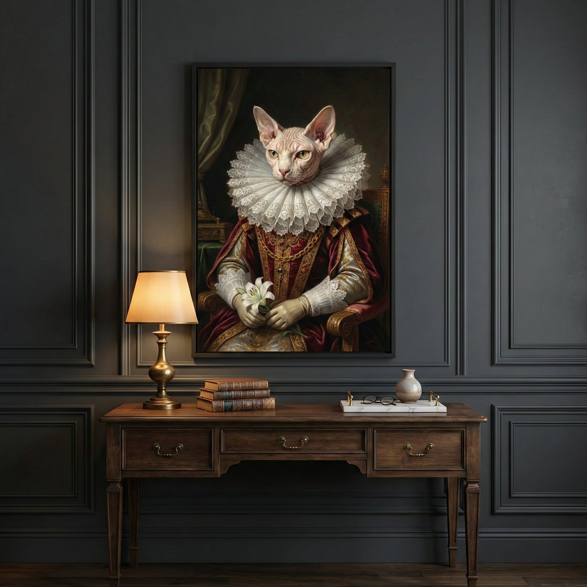 A framed portrait of a Sphynx cat dressed in regal historical attire, including a large white ruffled collar and an ornate red and gold robe. The cat holds a white lily in its paws. The artwork hangs on a dark paneled wall above a vintage-style wooden desk. A brass lamp with a warm glow, several leather-bound books, and a small decorative tray rest on the desk.