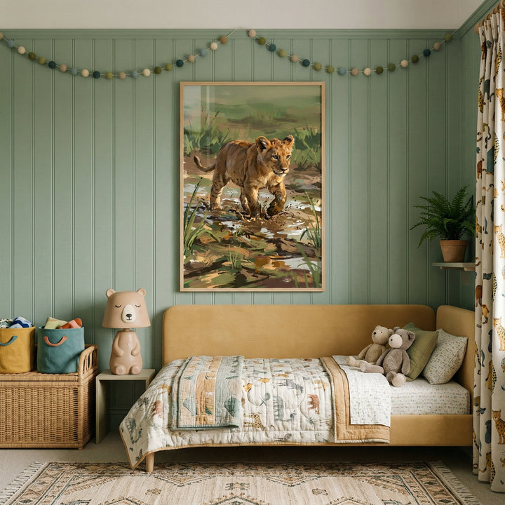 A child's bedroom with sage green paneled walls features a large framed illustration of a lion cub walking through a marsh. Above, a colorful pom-pom garland drapes across the wall. A mustard yellow upholstered daybed is dressed in animal-patterned bedding and holds several stuffed bears. Beside the bed, a bear-shaped lamp sits on a small table next to a woven storage chest with fabric bins. A patterned rug and curtains with animal motifs complete the cozy, safari-themed space.