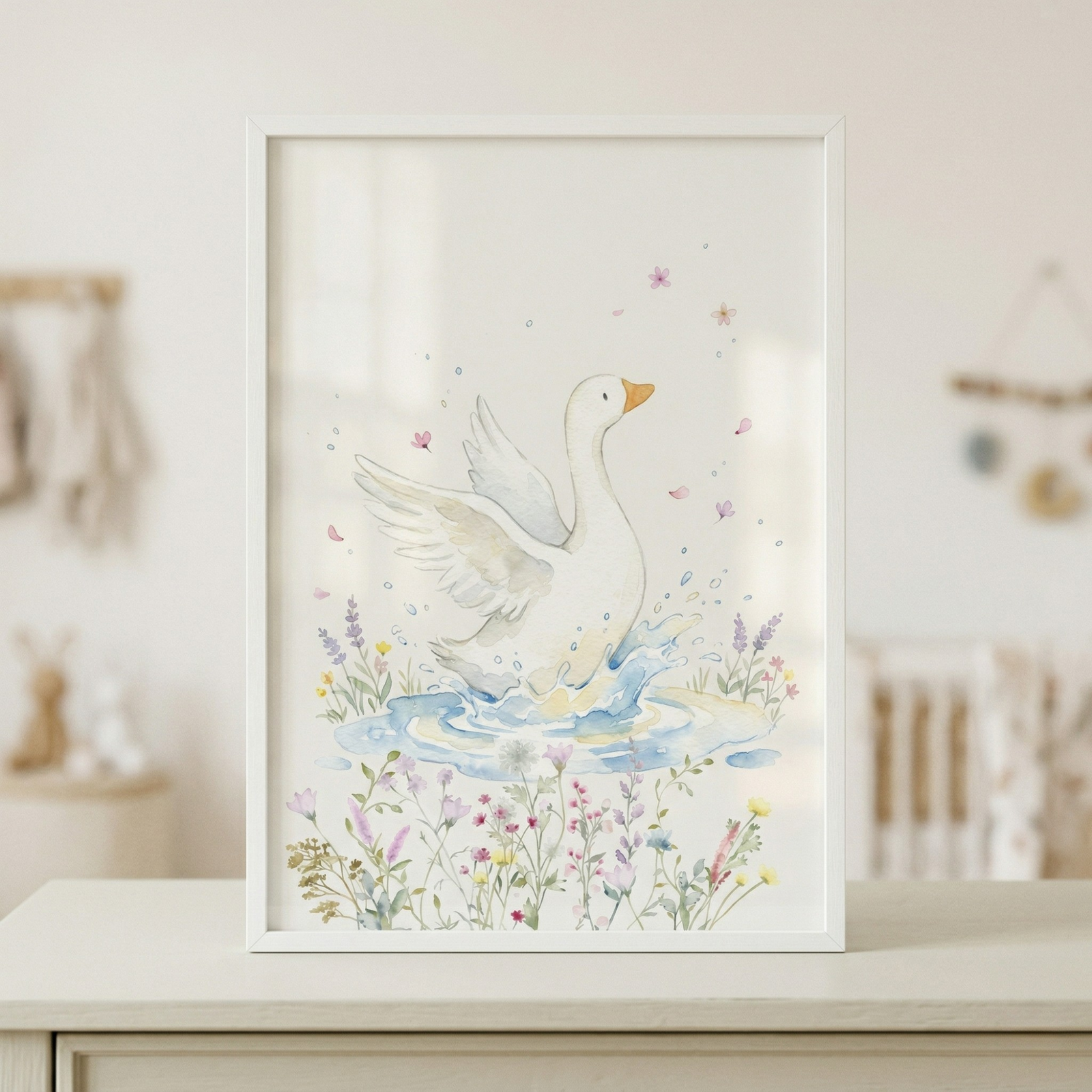 A framed watercolor-style illustration rests on a white dresser in a nursery. The artwork depicts a white goose splashing in a small blue pond with its wings spread. Delicate wildflowers in shades of pink, purple, and yellow bloom at the bottom, and tiny pink petals float in the air. The image is held in a thin white frame. The background is a softly blurred child's room featuring a crib and wooden wall decor, contributing to a gentle and peaceful setting.