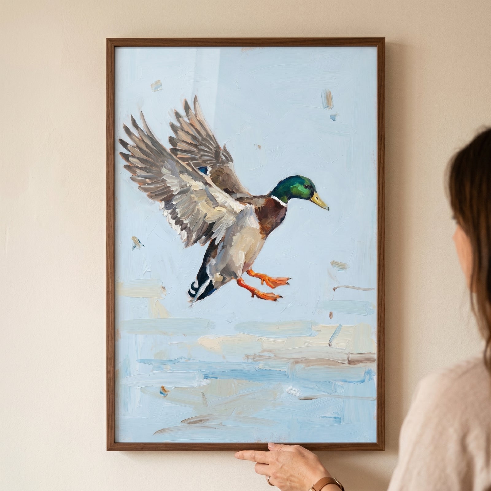 A person holds framed spread wing landing mallard wall art showing a duck with orange feet above light blue water.