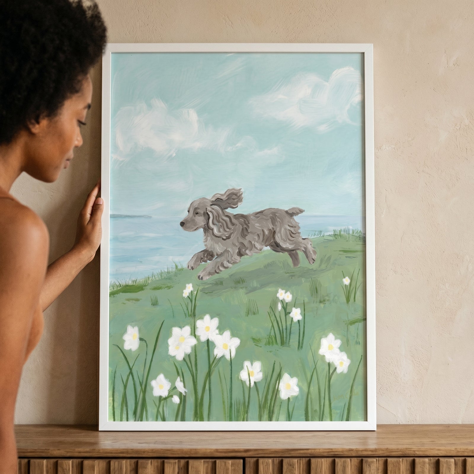 Woman holds spring meadow jumping spaniel dog wall art featuring a grey dog leaping through flowers near the coast.