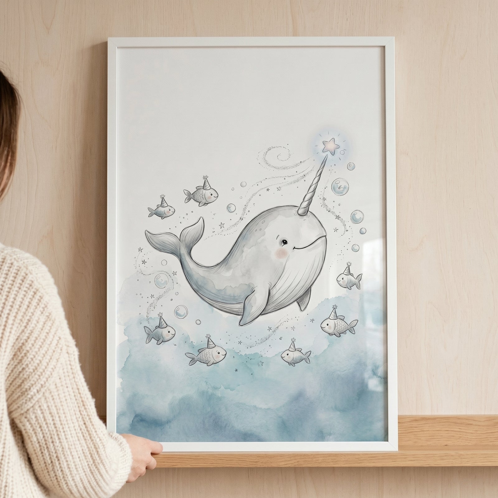 A person holds framed star tusk smiling narwhal wall art featuring whimsical fish in party hats and watercolor bubbles.