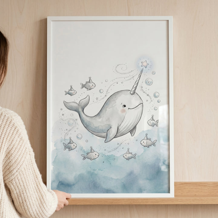 A person holds framed star tusk smiling narwhal wall art featuring whimsical fish in party hats and watercolor bubbles.