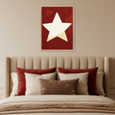 Modern bedroom with a beige upholstered bed. A framed artwork of a large white star with a golden glow on a textured red background hangs above the bed. The bed has red, cream, and brown pillows, including one with gold constellation embroidery.