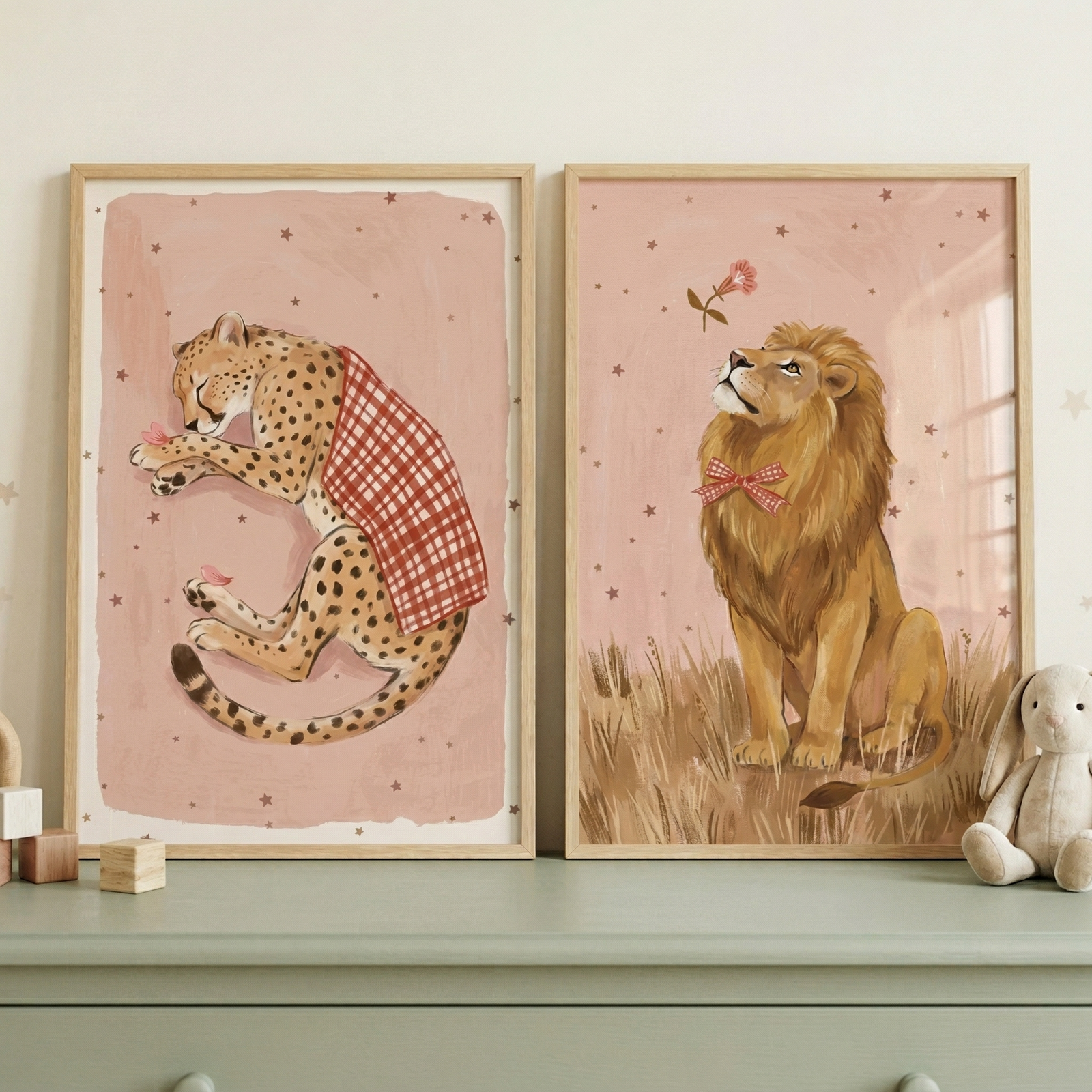 Two framed animal illustrations sit on a shelf styled with wooden toys and a plush bunny. One shows a spotted cheetah curled up wearing a red checkered cloth and holding a flower, while the other shows a lion with a full mane and matching bow tie sitting in grass and looking up at a floating flower against a soft pink star patterned background.