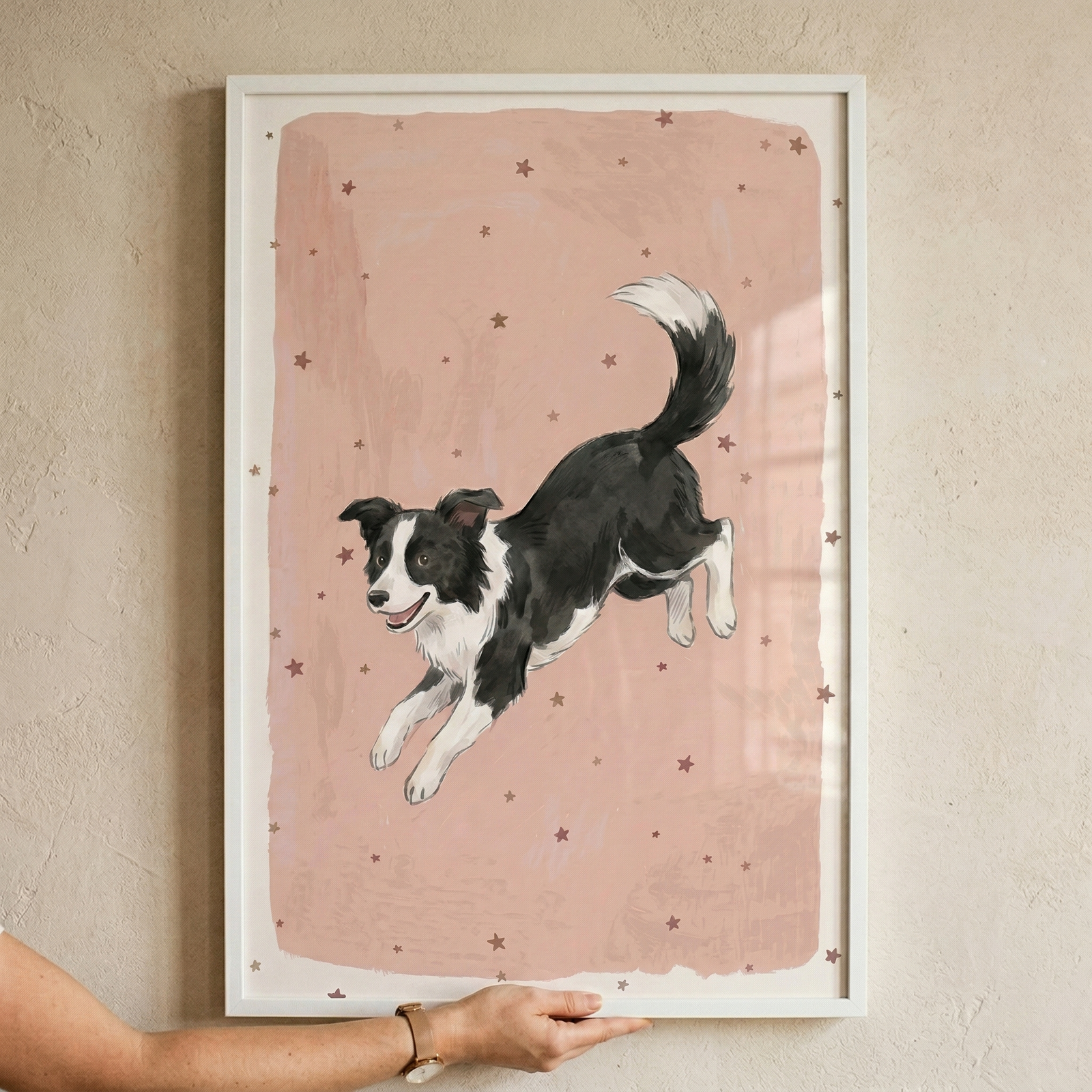A framed illustration of a black and white Border Collie leaping through the air. The dog is shown in a dynamic pose with its front paws reaching forward and its tail curved gracefully upwards. It has an energetic and happy expression. The background is a textured, soft pink rectangle with uneven edges, decorated with small, dark star shapes. The artwork is held in a minimalist white frame by a person whose hand and arm are visible at the bottom. 