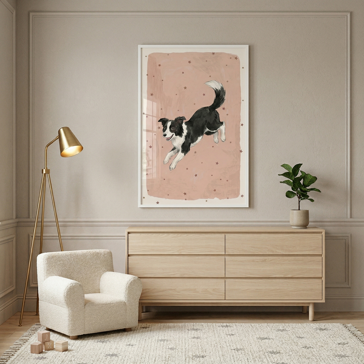 A photograph of a modern room interior featuring neutral tones and soft decor. Centered on a light beige wall with decorative molding is a white-framed vertical artwork. The illustration depicts a black and white border collie jumping through the air against a muted pink background scattered with small stars.

