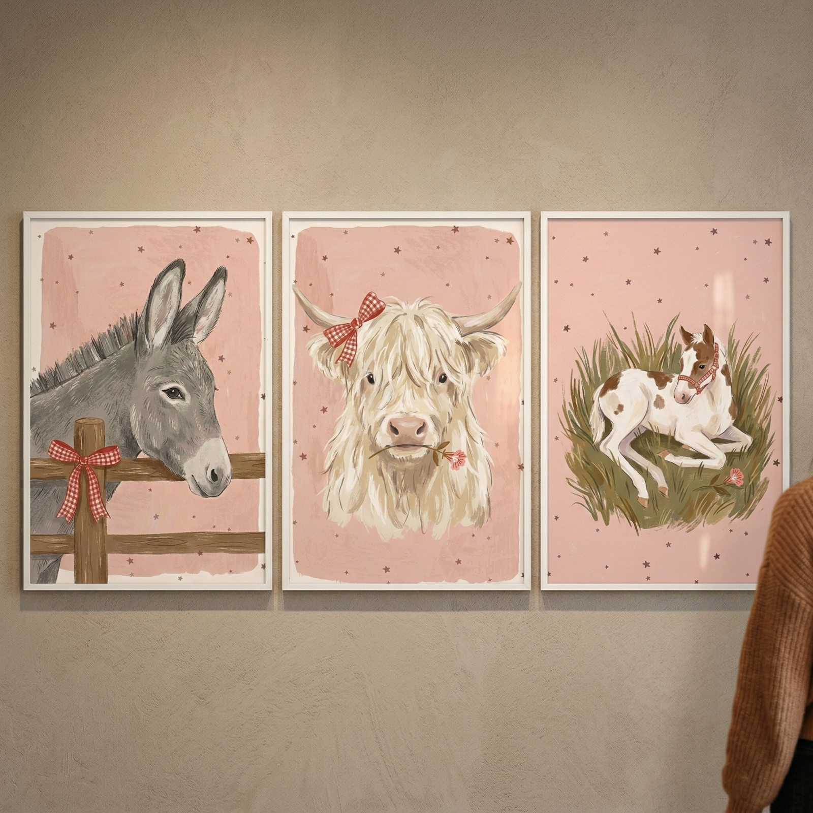 Three white-framed art pieces hang side-by-side on a plain wall. Each piece has a soft pink background dotted with small stars. The left illustration features a grey donkey looking over a wooden fence with a red checkered bow. The middle piece shows a fluffy tan cow with a red checkered bow on its horn and a small pink flower in its mouth. The right piece depicts a spotted brown and white foal lying in green grass. Part of a person in a brown sweater is visible on the right.