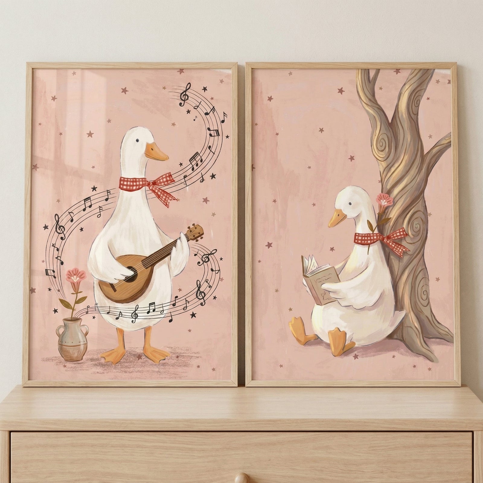 Two framed pastel illustrations show a white duck wearing a red gingham bow tie, one playing a guitar with musical notes swirling around it and the other sitting under a tree reading a book. The soft pink background and cozy scenes create a calm and imaginative nursery wall art set.