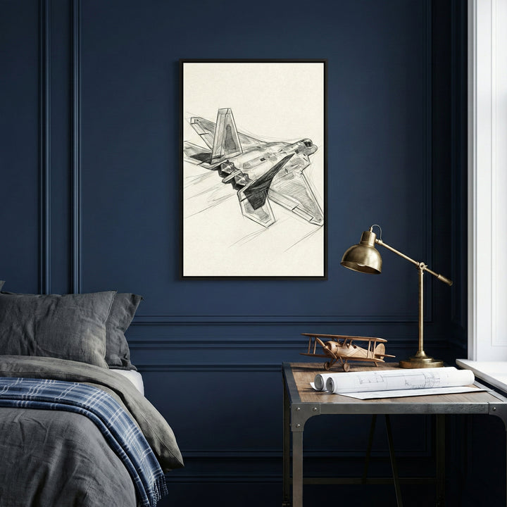 A framed technical sketch of a military jet fighter hangs on a dark navy blue wall with decorative molding. The sketch shows the aircraft from a rear-three-quarter perspective. Below the artwork, a wooden desk with industrial metal legs is positioned against the wall, holding a brass lamp, a small wooden model airplane, and rolled-up blueprints. To the left, part of a bed with grey pillows and a blue-and-grey plaid blanket is visible. Natural light comes from a window on the right.