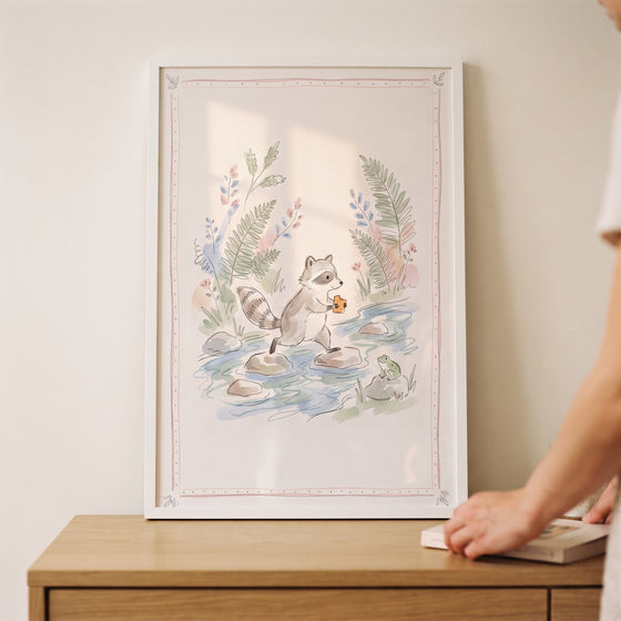 Framed stepping stone raccoon wall art showing a watercolor raccoon crossing a creek on rocks while holding a snack.