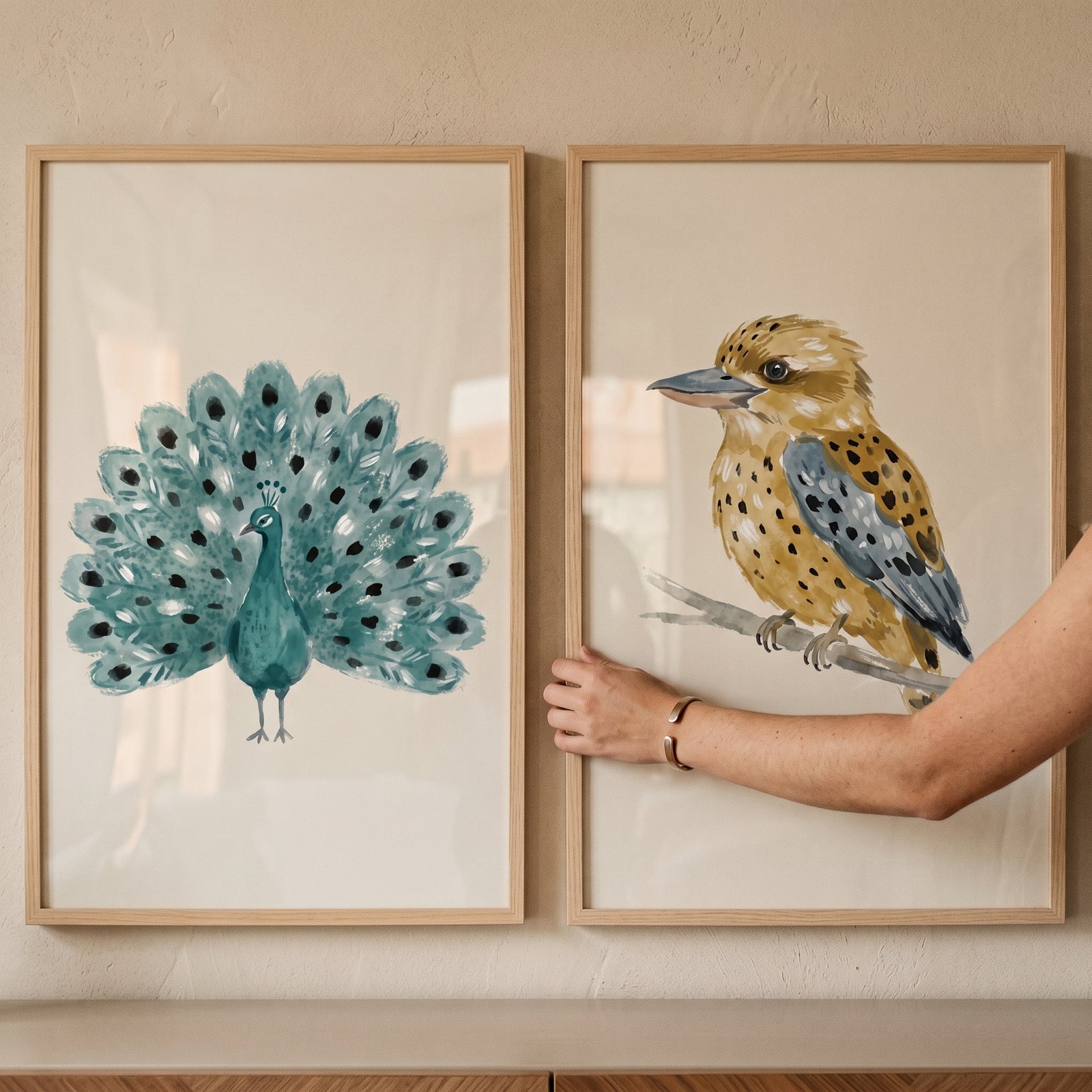 A hand touches a stippled earth tone birds wall art set of 2 featuring a teal peacock and a tan and blue bird.