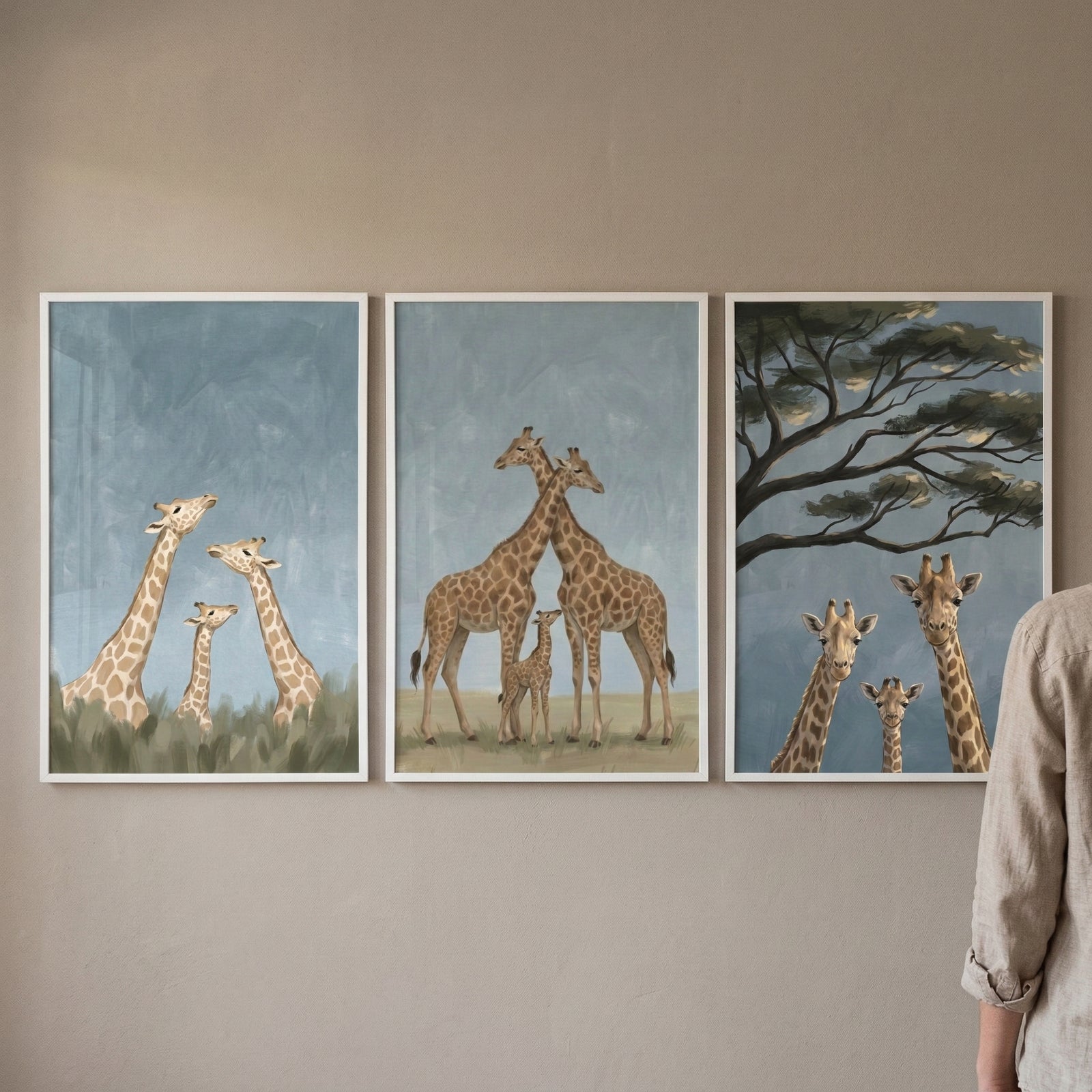 Three framed giraffe prints hang in a row on a taupe wall with a person standing at the right edge for scale. The artwork shows giraffe families in soft blue and green landscapes, including one scene with tall grass, one with two adults and a calf, and one beneath spreading tree branches.