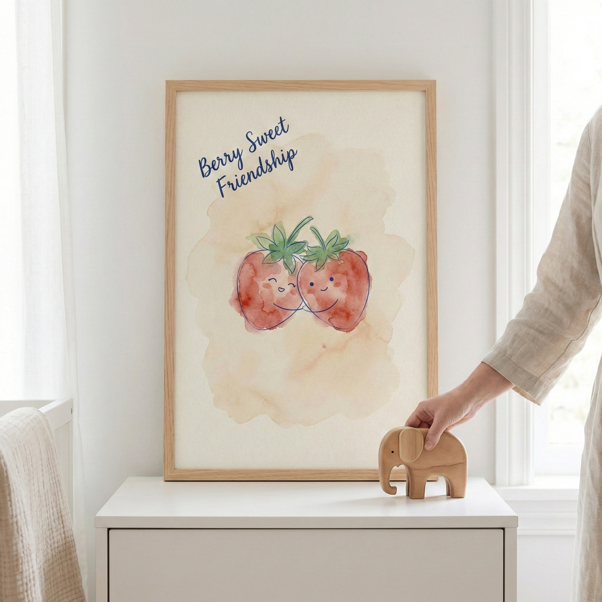 A light-colored wooden frame sits atop a white dresser, displaying a piece of art with a soft watercolor-style illustration. The artwork features two smiling strawberries embracing against a warm, hazy background. Above the characters, the words "Berry Sweet Friendship" are written in a dark blue script. To the right, a person’s hand is placing a simple, carved wooden elephant toy on the surface next to the frame. 