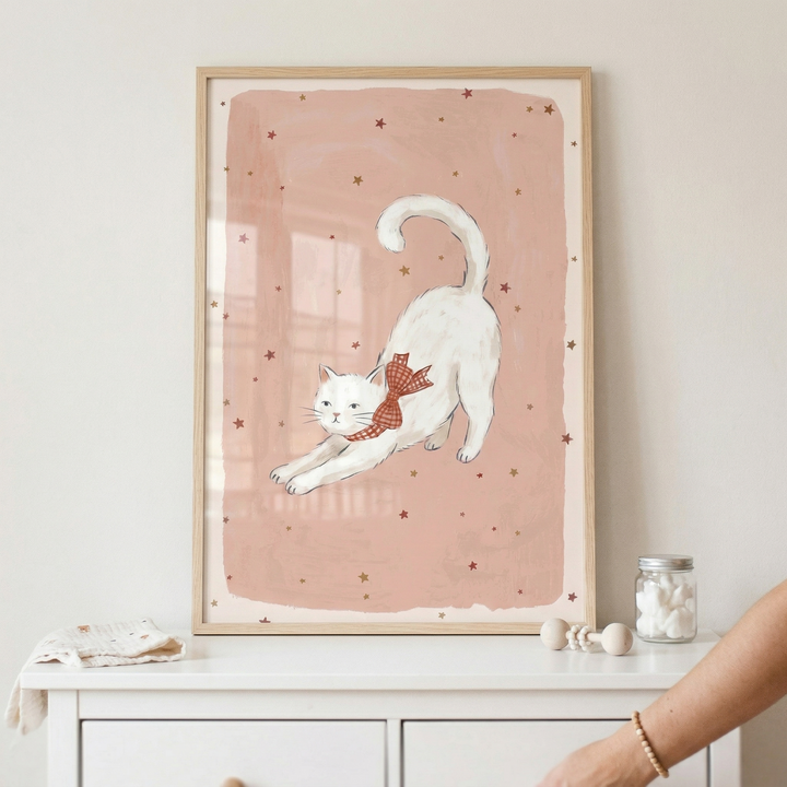 A framed illustration of a white cat stretching is displayed on a light-colored wall above a white wooden dresser. The cat is shown in a playful stretch, wearing a red and white checkered bow around its neck. The artwork features a soft pink rectangular background decorated with small, scattered brown and gold stars. The piece is encased in a simple, light-toned wood frame. 