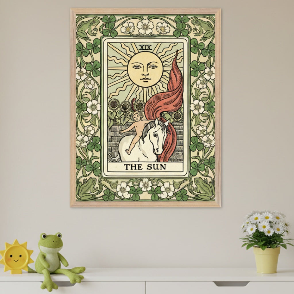 Kid's room with a white dresser. A framed print of "The Sun" tarot card hangs centered above the dresser. The card depicts a sun, a child on a white horse, and sunflowers, framed by an art nouveau border with foliage and frogs. A sun toy, frog plush, and potted daisies are on the dresser.