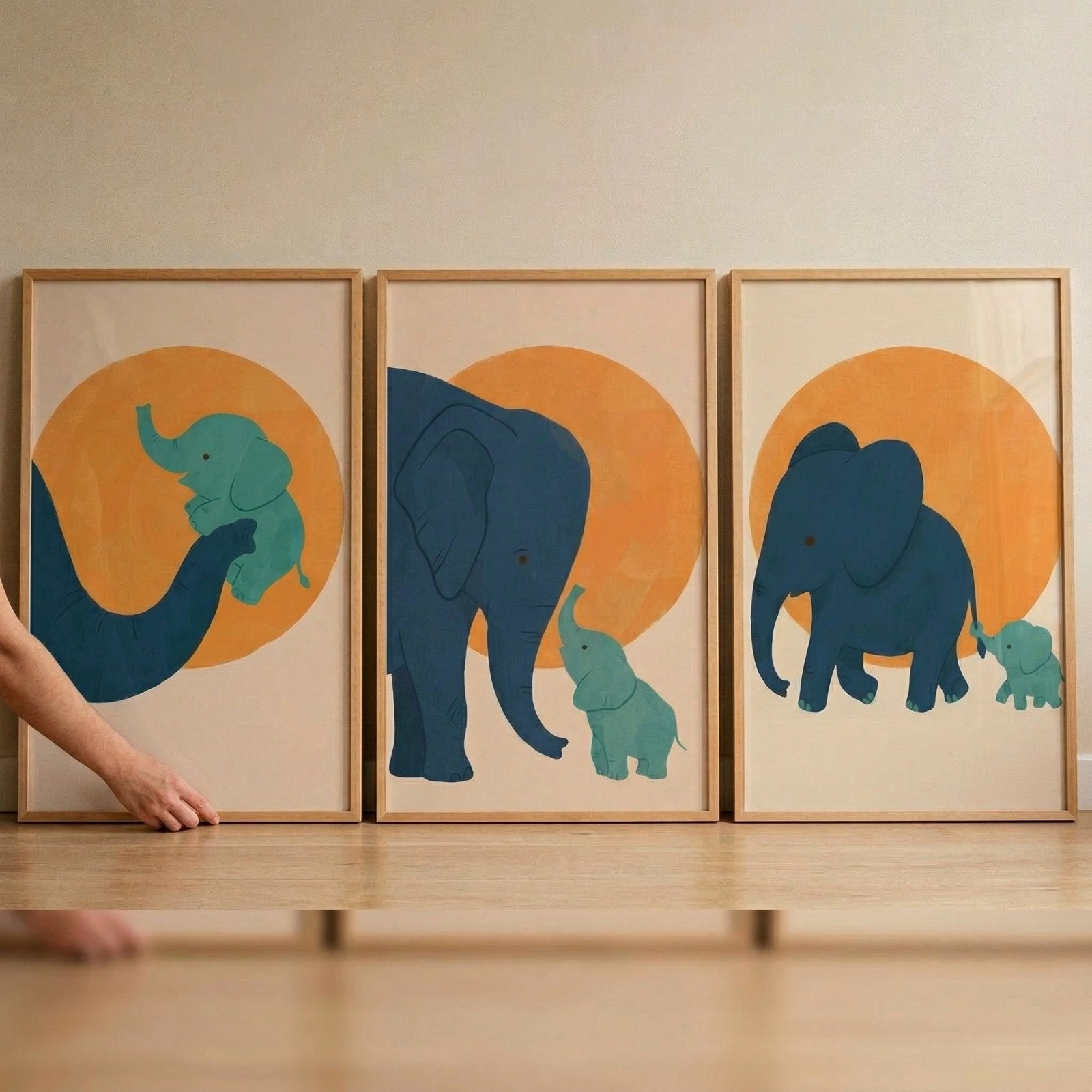 A set of three framed minimalist illustrations featuring elephants is displayed on a light wood floor against a plain, neutral wall. Each piece of art is housed in a light wood frame and features a large orange circle in the background. 