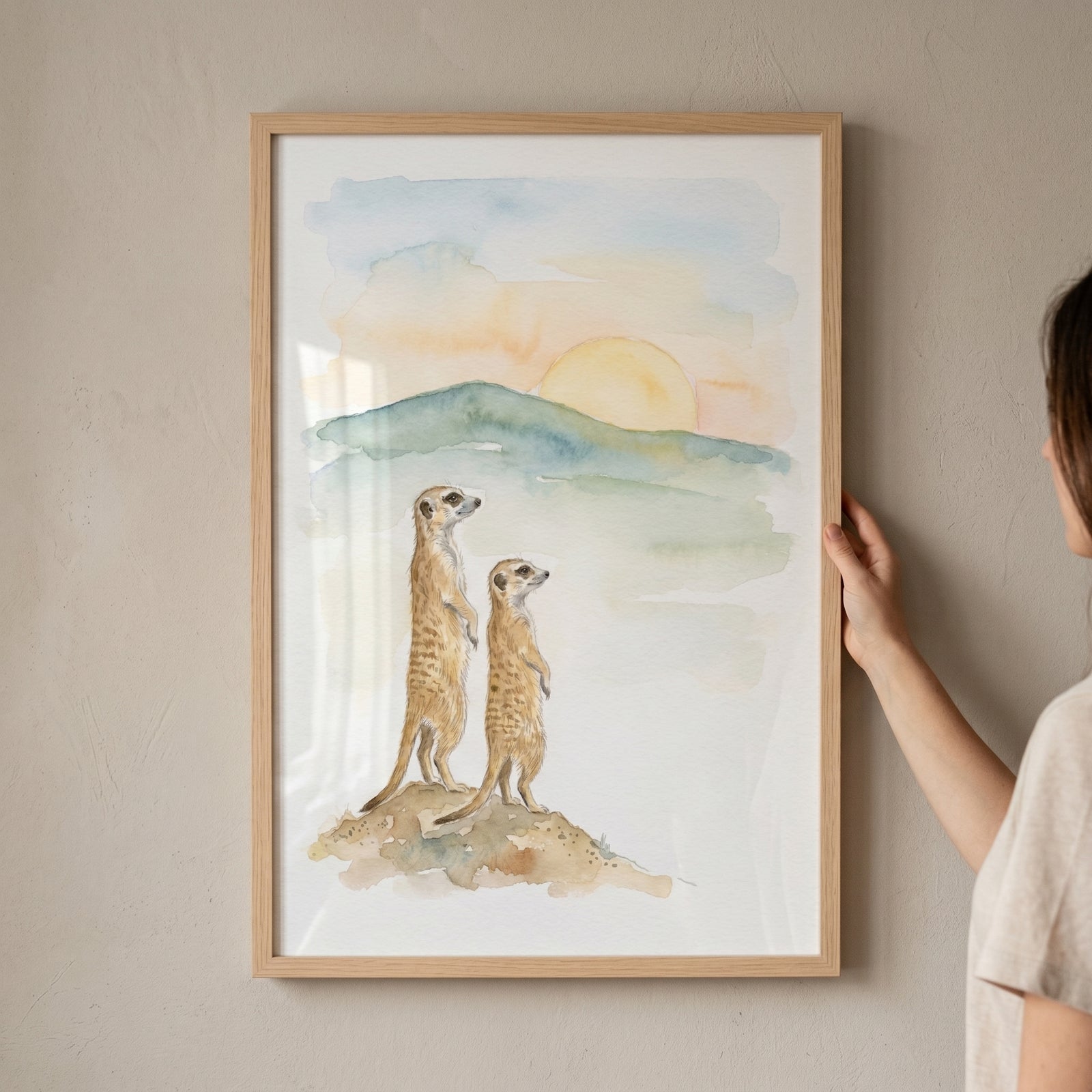 Watercolor sunset meerkat wall art of two meerkats standing on a hill with a sun setting behind distant mountains.