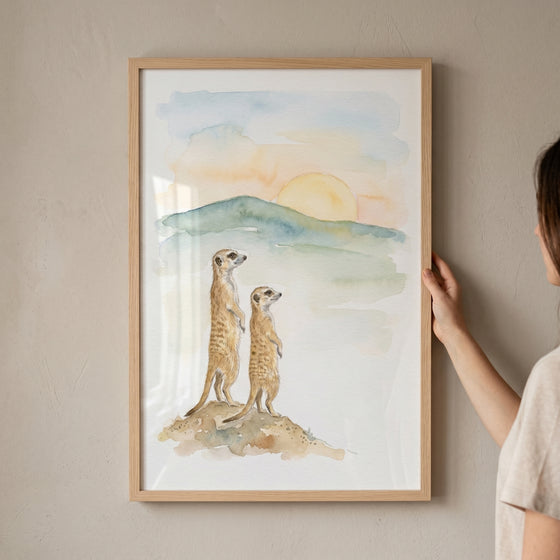 Watercolor sunset meerkat wall art of two meerkats standing on a hill with a sun setting behind distant mountains.