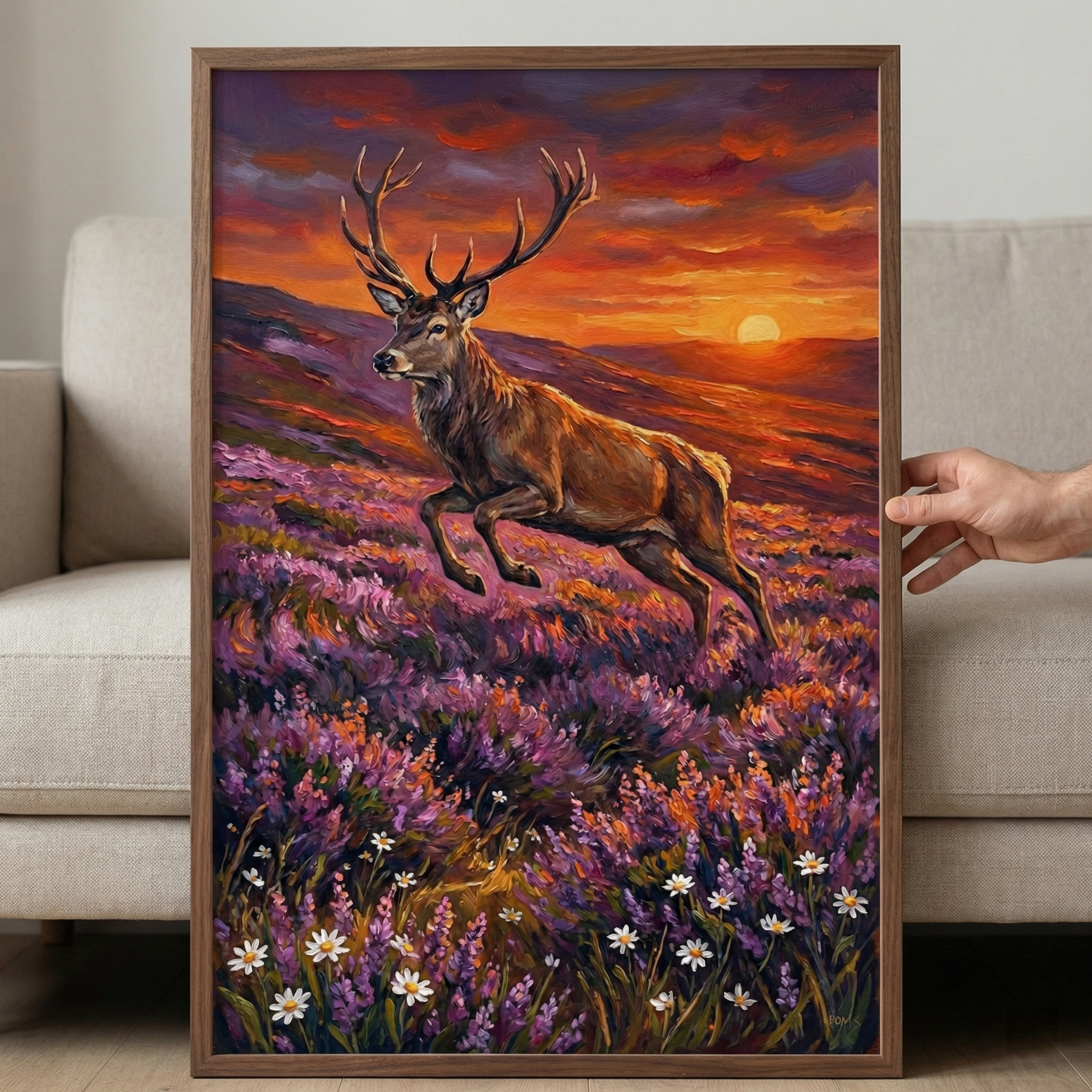 A vertical framed artwork held by a hand depicts a majestic stag leaping over a field of purple heather and white daisies. The scene is set against a vivid sunset, with the sun low on the horizon behind rolling hills. The sky is filled with dramatic clouds in shades of orange, red, and deep purple. The artwork has a textured, expressive style. The frame is a dark wood, and the piece is held up against a light-colored sofa.