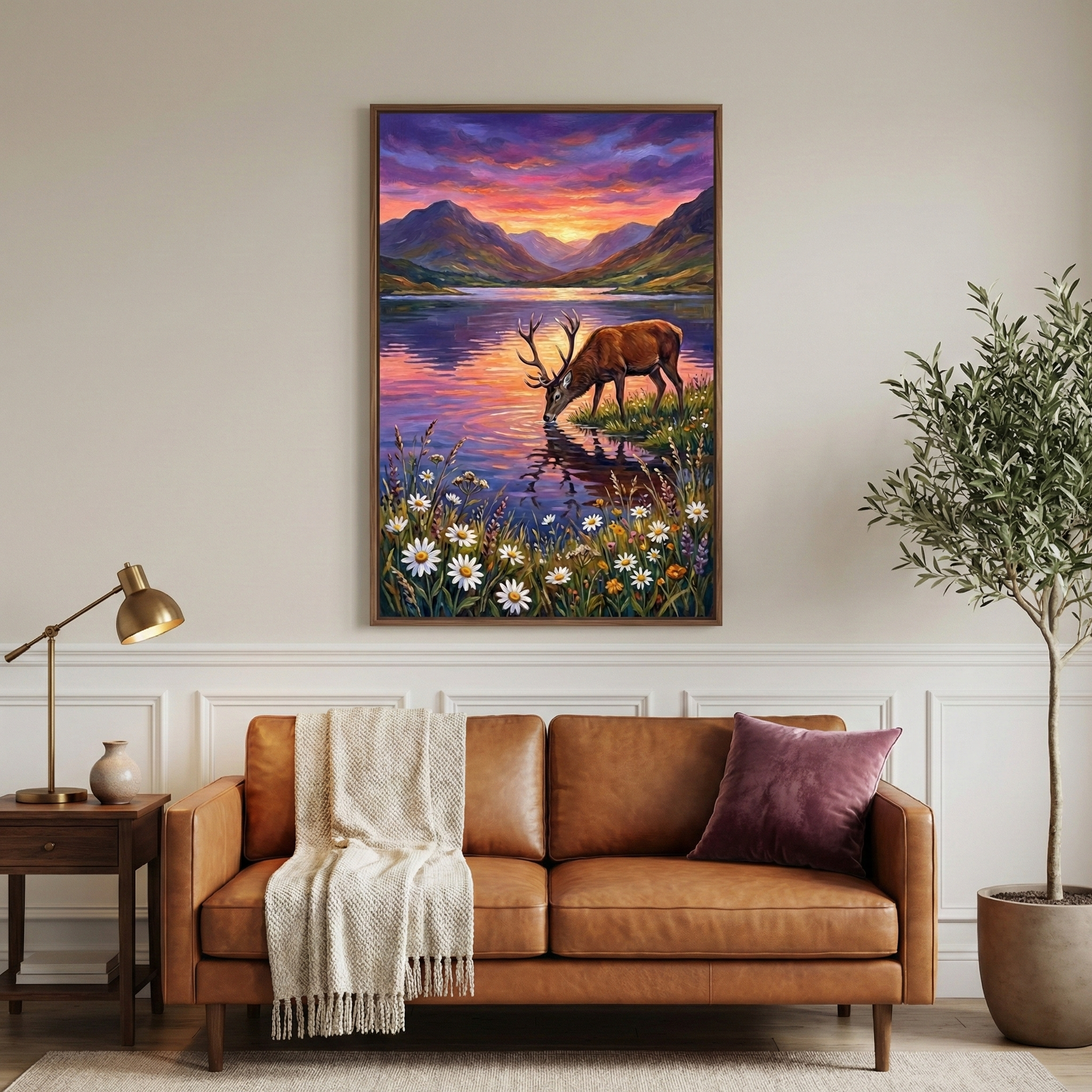 "A vertically oriented piece of artwork is displayed centrally on a neutral, paneled wall above a modern living room setup. The artwork depicts a majestic stag with large antlers drinking from a tranquil lake at sunset. The sky is a vibrant gradient of purple, pink, and orange, reflected beautifully in the calm water. Rolling mountains are silhouetted against the horizon. In the immediate foreground of the image, a lush cluster of white daisies and yellow wildflowers grows along the bank.

