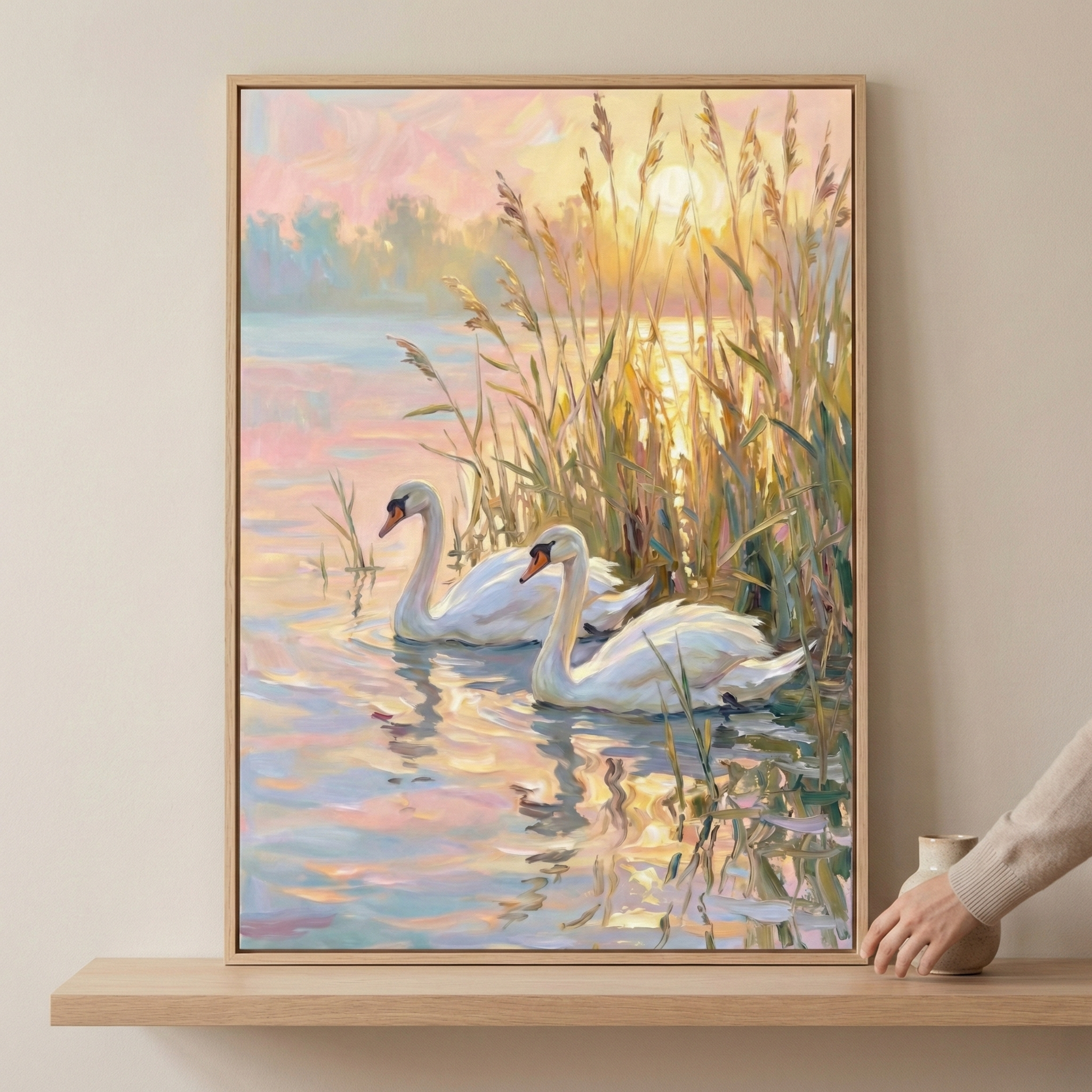A framed painting of two elegant swans gliding on a tranquil lake, surrounded by tall reeds, with the warm glow of the sunset reflecting on the water. A hand is adjusting the artwork on a wooden shelf.