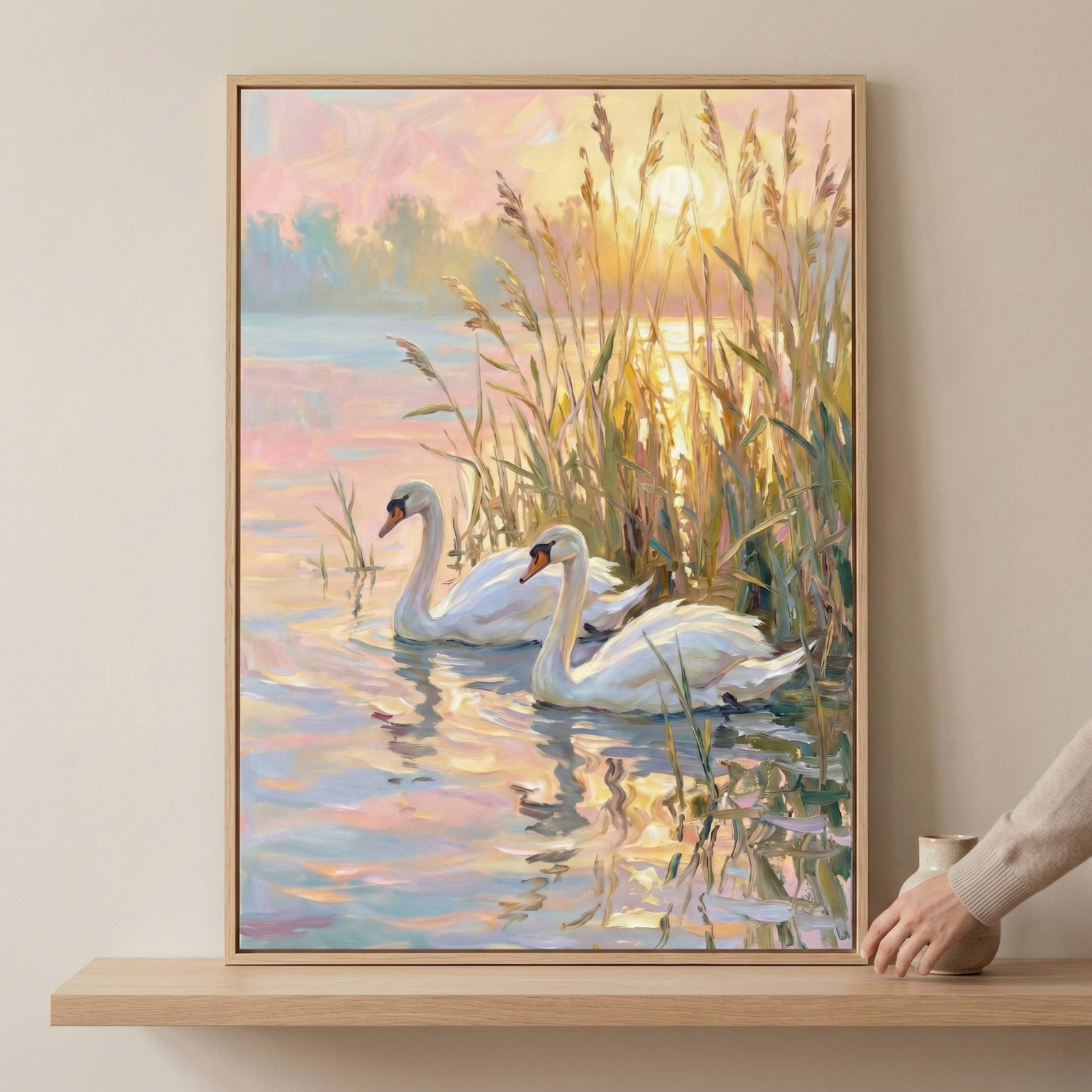 A framed painting of two elegant swans gliding on a tranquil lake, surrounded by tall reeds, with the warm glow of the sunset reflecting on the water. A hand is adjusting the artwork on a wooden shelf.
