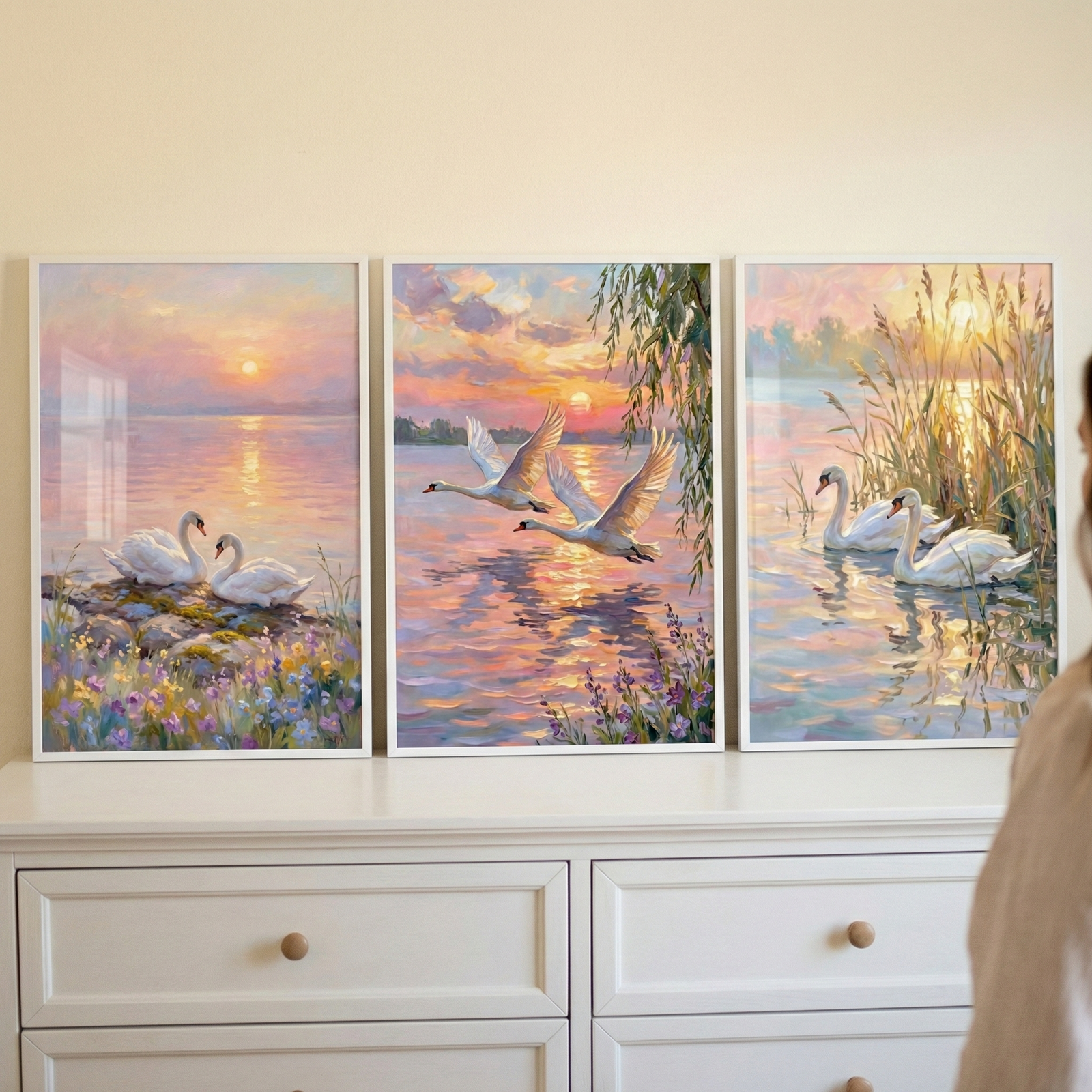 Three framed art pieces are arranged in a row on top of a white wooden dresser. Each piece features an impressionistic depiction of white swans at sunset in soft shades of pink, orange, and purple. The leftmost piece shows two swans resting on a flowery bank by a lake. The center piece depicts two swans in flight over shimmering water with a willow branch in the corner. The rightmost piece shows two swans swimming among tall golden reeds. 
