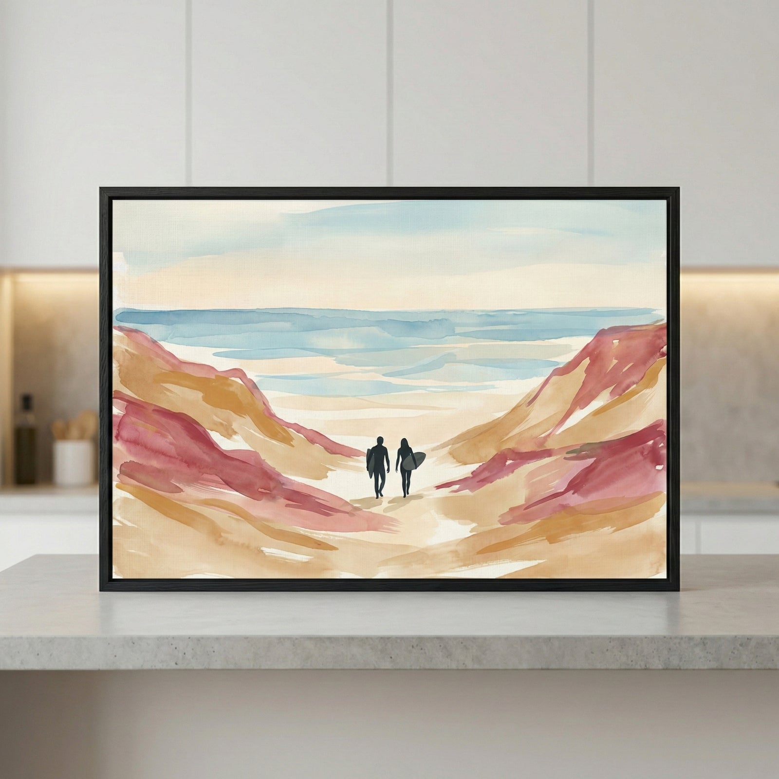 A horizontal, framed watercolor-style illustration sits on a light-colored countertop. The artwork depicts two dark silhouetted figures carrying surfboards, walking between tall, abstract sand dunes toward a calm sea. The dunes are rendered in soft washes of tan, ochre, and muted reddish-pink. The water and sky transition from pale cream to light blue. The piece is enclosed in a simple black frame.