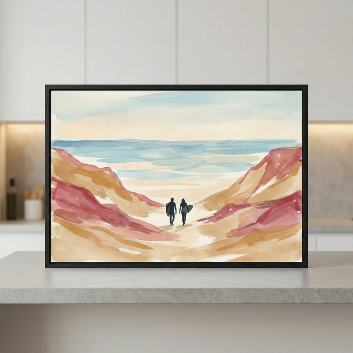 A horizontal, framed watercolor-style illustration sits on a light-colored countertop. The artwork depicts two dark silhouetted figures carrying surfboards, walking between tall, abstract sand dunes toward a calm sea. The dunes are rendered in soft washes of tan, ochre, and muted reddish-pink. The water and sky transition from pale cream to light blue. The piece is enclosed in a simple black frame.