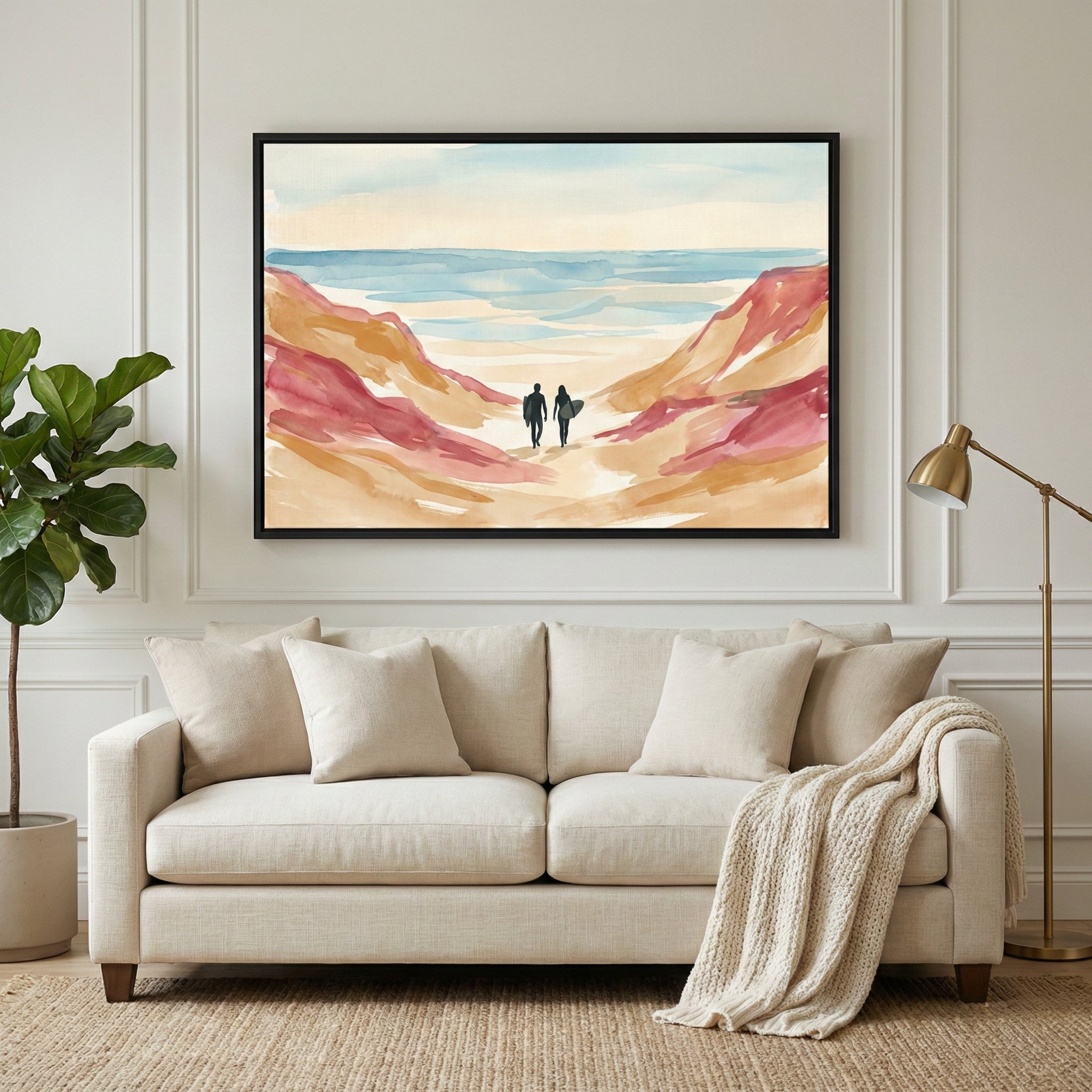 A minimalist living room features a cream-colored sofa with several matching pillows and a textured throw blanket. Above the sofa hangs a large framed horizontal artwork depicting two small silhouetted figures walking through flowing, watercolor-style dunes toward a serene blue ocean. The room has elegant white paneled walls, a large green leafy plant in a simple pot to the left, and a brass floor lamp to the right. A neutral-toned woven rug covers the floor.