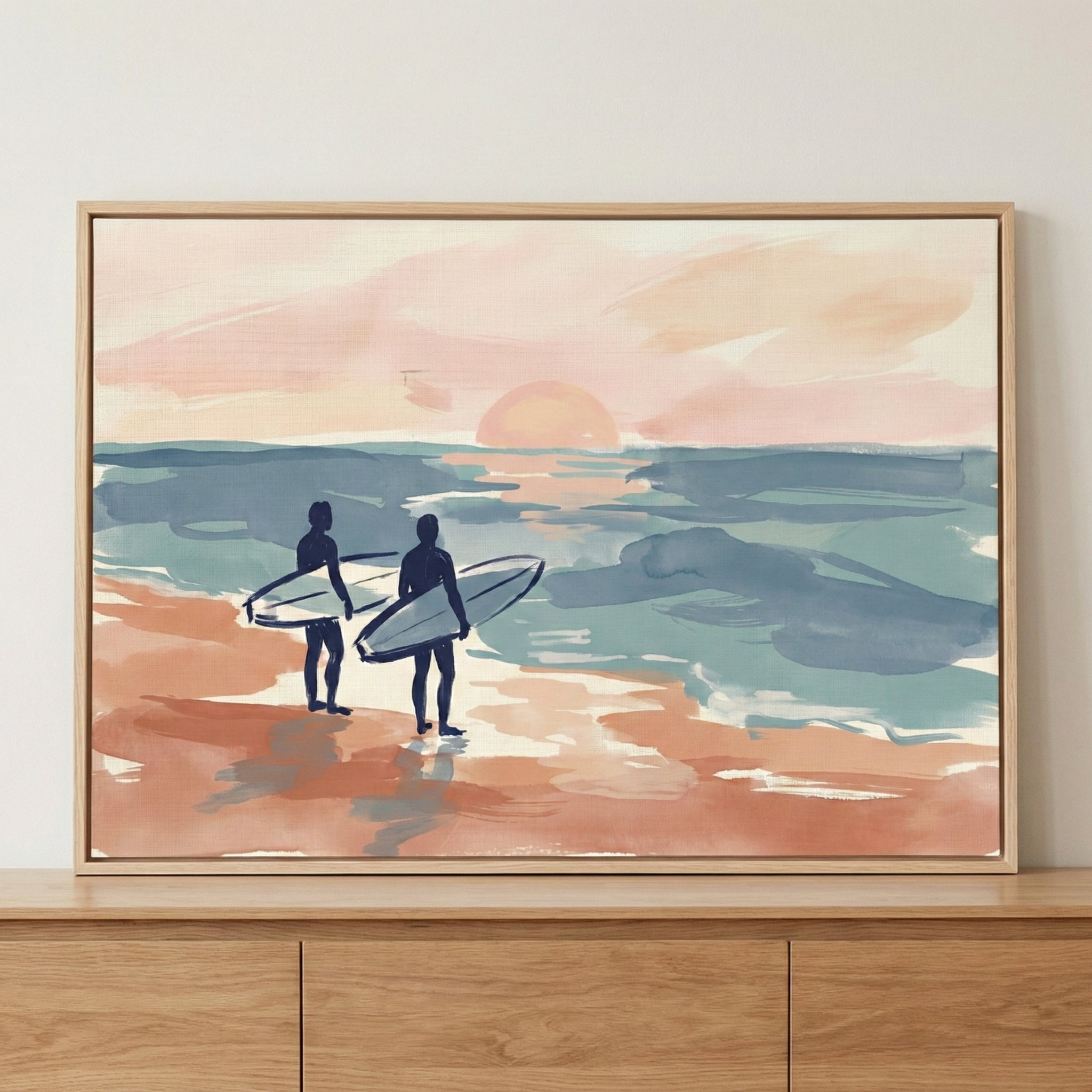 A framed illustration of two dark-blue silhouettes of surfers standing on a peach-colored beach, holding their surfboards and looking out at a calm, teal and blue ocean. A large, soft-peach sun sits low on the horizon in a pale pink sky. The style is soft and expressive with visible brushstrokes and a muted pastel color palette.