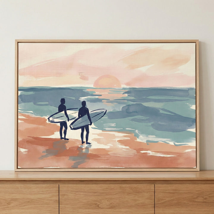 A framed illustration of two dark-blue silhouettes of surfers standing on a peach-colored beach, holding their surfboards and looking out at a calm, teal and blue ocean. A large, soft-peach sun sits low on the horizon in a pale pink sky. The style is soft and expressive with visible brushstrokes and a muted pastel color palette.