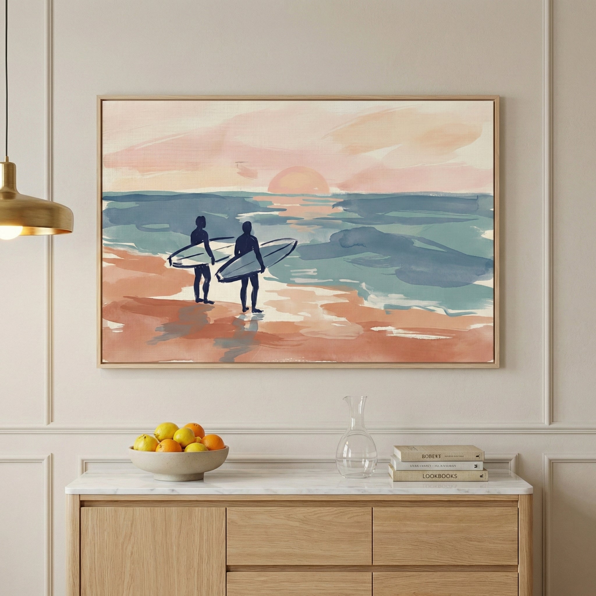 A large framed horizontal piece of artwork hangs on a light-colored wall with minimalist molding. The image within the frame shows two silhouetted figures carrying surfboards as they walk along a beach toward a calm ocean. A pale sun sits low on the horizon, casting a soft glow across the sky and water. The scene is rendered with broad, horizontal strokes in a color palette of peach, teal, and terracotta. 