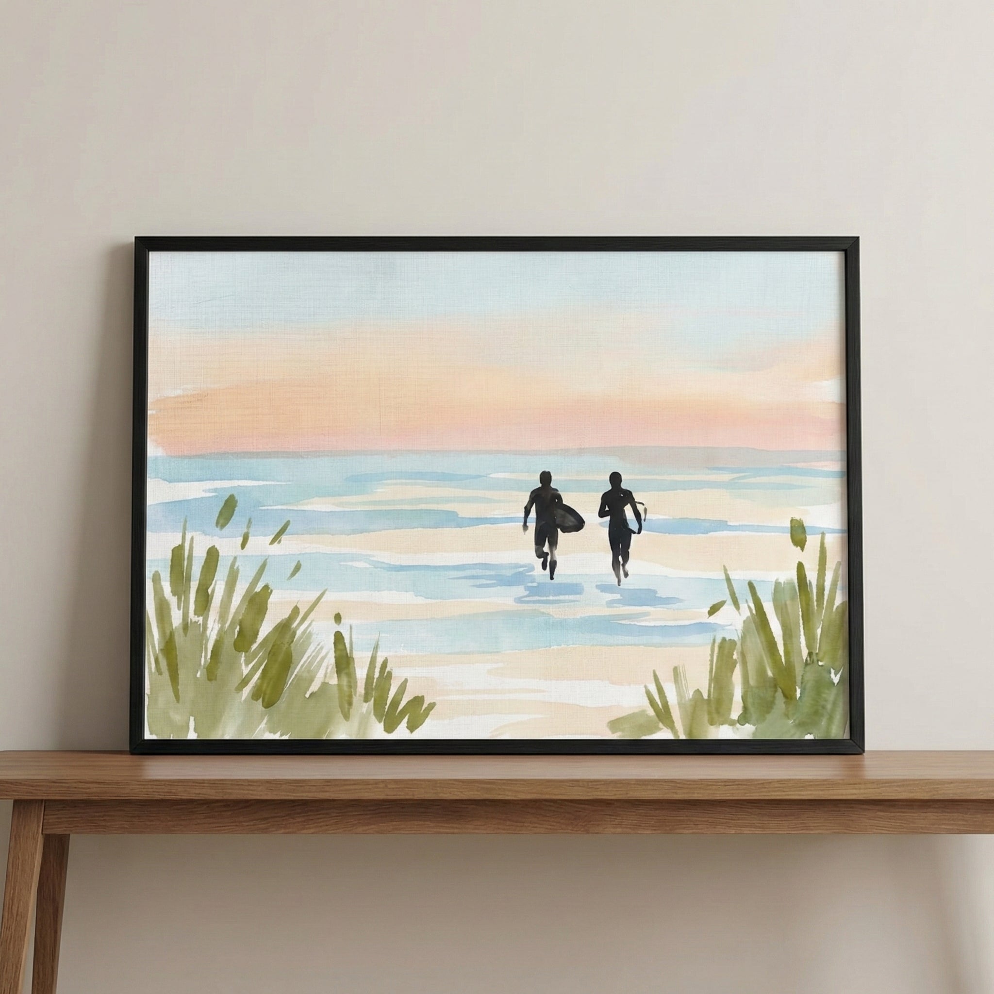 A framed landscape artwork depicting a serene beach scene at sunset. Two dark silhouettes of figures, one holding a surfboard, are walking through shallow water toward the horizon. The sky is rendered in soft washes of peach, cream, and light blue, mirrored by the gentle waves below. In the foreground, tufts of green coastal grass frame the bottom corners of the composition. The piece is set within a thin black frame and sits on a wooden console table against a neutral wall.