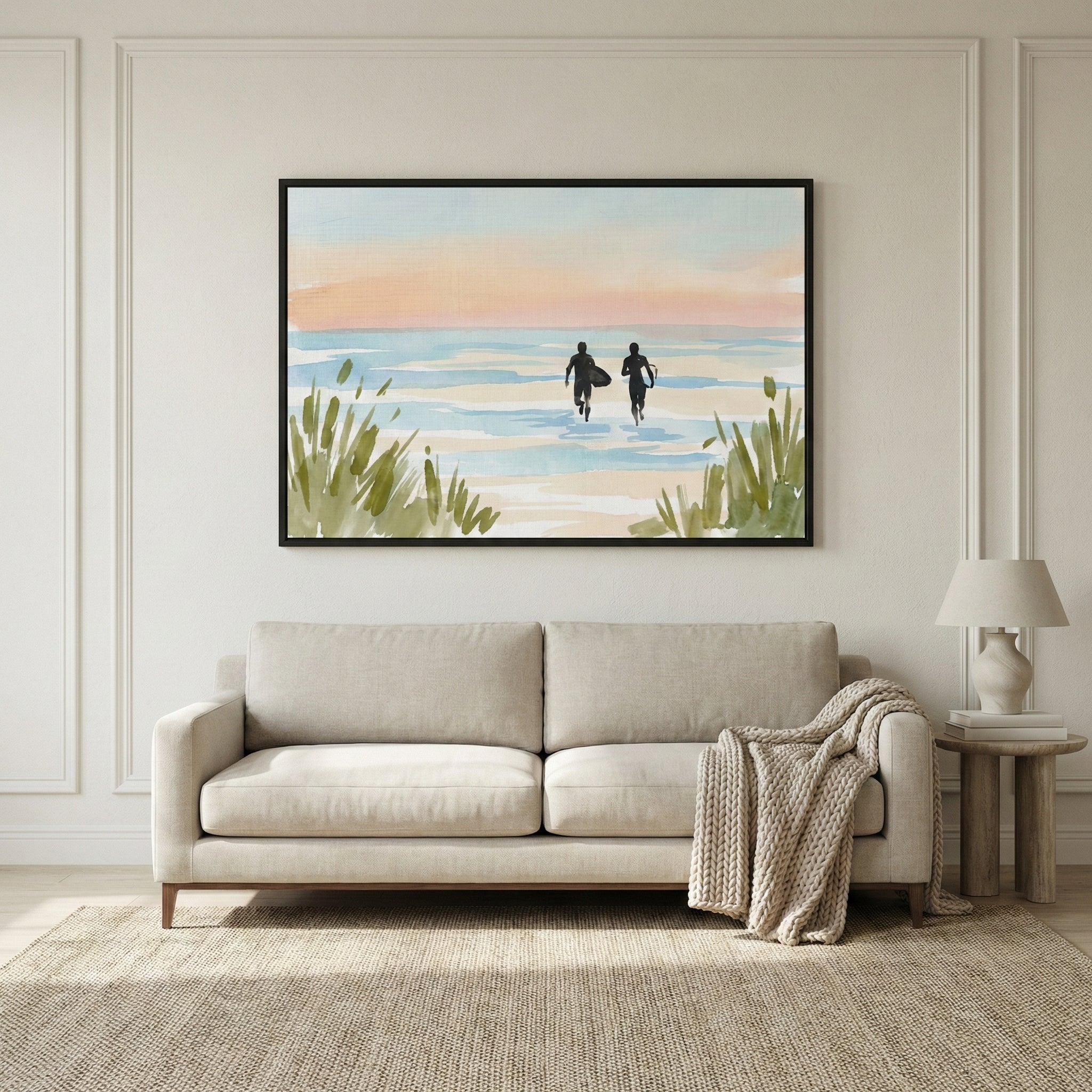 A minimalist watercolor artwork framed and mounted on a neutral wall above a light beige sofa. The piece shows two dark silhouettes of surfers walking into a pale blue sea under a soft peach and pink sky, framed by green coastal grass in the foreground. The room is decorated with a textured rug and a wooden side table holding a cream lamp and books.