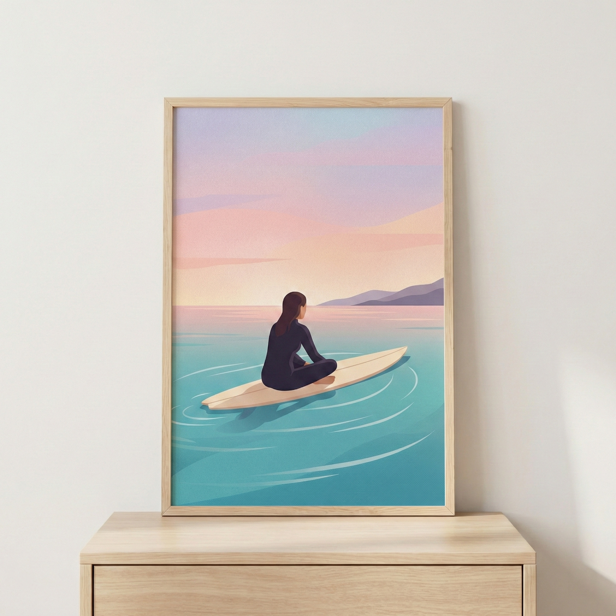 A minimalist art print in a light wood frame rests on a wooden surface against a neutral wall. The illustration depicts a person with dark hair wearing a black wetsuit, sitting cross-legged on a light-colored surfboard. The figure is viewed from behind, looking out over a calm turquoise ocean toward distant purple mountains. The sky above is filled with soft horizontal bands of pastel pink, peach, and blue, suggesting a quiet sunrise or sunset. Subtle white ripples surround the surfboard in the still water.