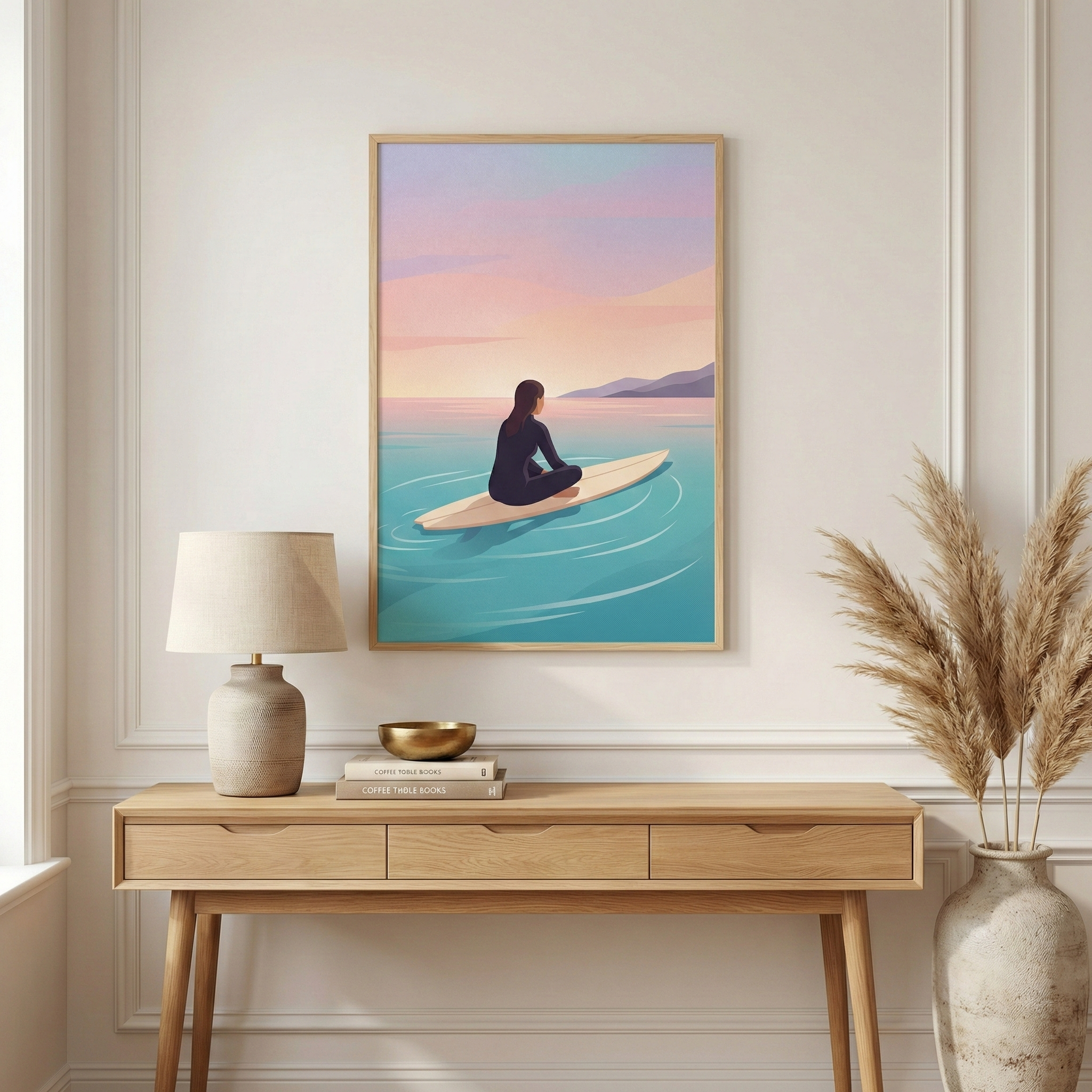 A vertical framed illustration hangs on a white wall above a wooden console table. The artwork shows a person in a dark wetsuit sitting on a surfboard in a calm ocean under a pastel sunset sky with shades of pink and orange. On the light-colored wooden table below, there is a lamp with a textured base and a cream shade, a stack of books with a small golden bowl on top, and a large ceramic vase filled with dried grass on the right. The room features a clean, minimalist aesthetic with soft, natural lighting.