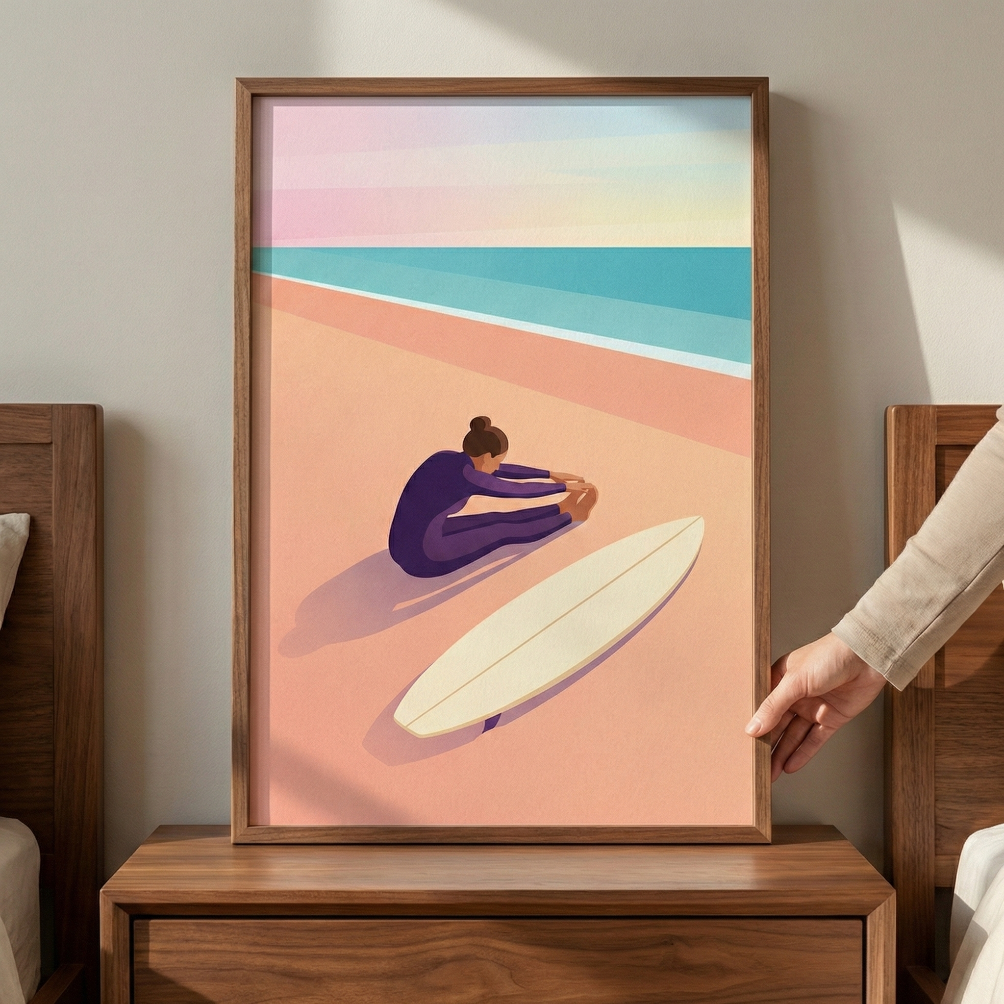 A framed minimalist illustration of a person in a purple suit stretching on a peach-colored beach next to a white surfboard. Behind the figure, a multi-toned blue ocean meets a soft pastel sky with horizontal bands of pink, yellow, and light blue. The wood-framed artwork rests on a dark wood nightstand between two headboards, and a hand is seen touching the right side of the frame.