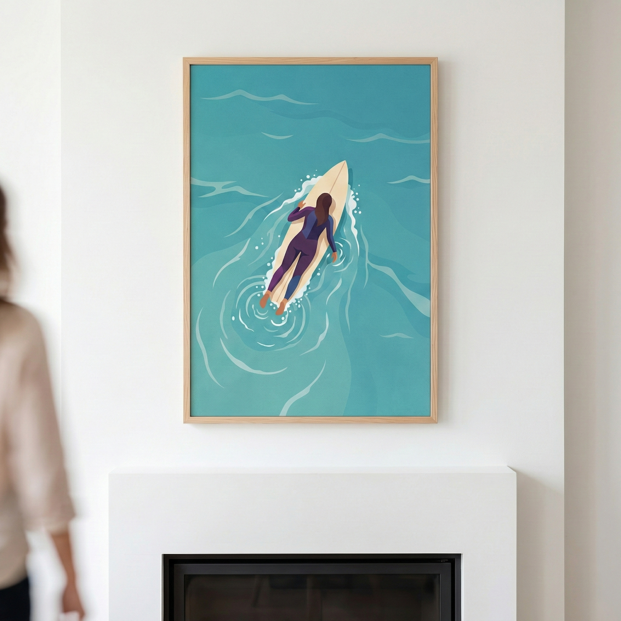 A framed illustration hangs on a white wall above a modern white fireplace. The vertical piece depicts an overhead view of a woman lying on a light-colored surfboard in turquoise water. She wears a dark purple wetsuit and is surrounded by soft ripples and small trails of white foam. The artwork is set in a light-toned wooden frame. To the left, a blurred figure of a person is partially visible, walking past the display.