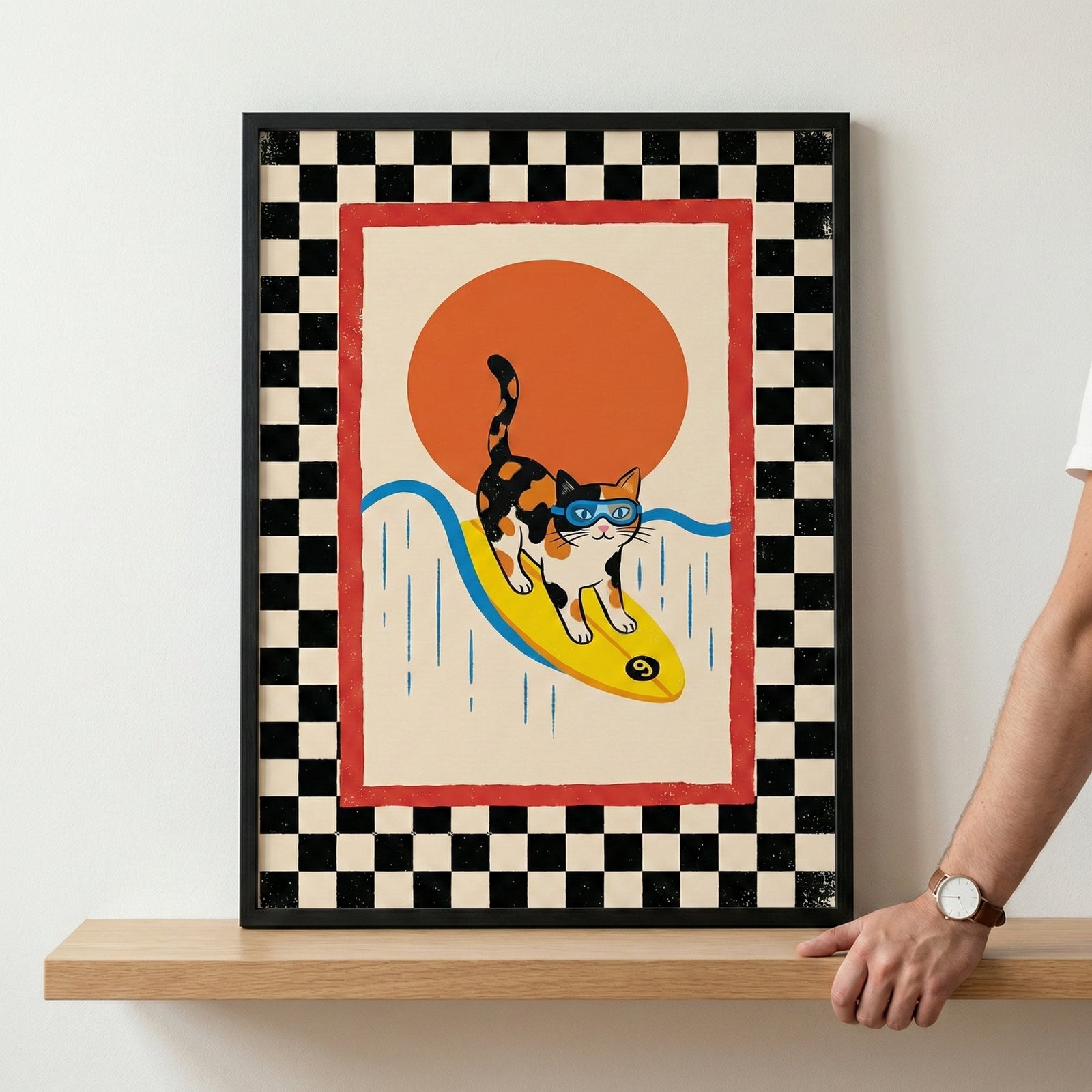 A framed graphic illustration sits on a light-colored wooden shelf against a neutral wall. The artwork depicts a tricolored calico cat wearing blue goggles while riding a yellow surfboard. Behind the cat is a large orange circular sun and stylized blue wavy lines. The central image is enclosed within a solid red border, which is itself surrounded by a wide black and white checkered pattern. A person's arm and hand, wearing a watch, are visible on the right side of the frame, resting on the shelf.