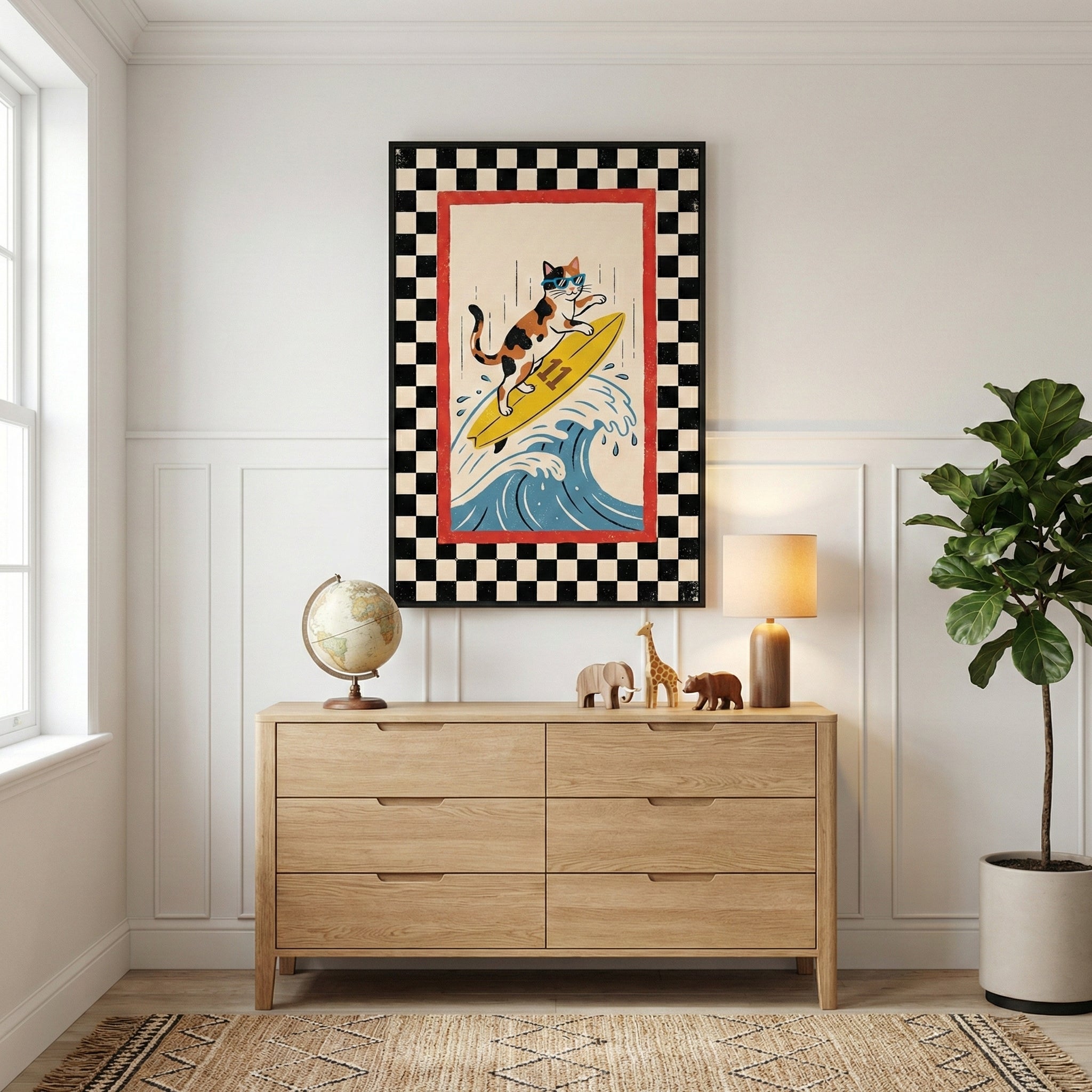 A piece of framed wall art hangs on a white wall above a light wood dresser. The illustration features a calico cat wearing sunglasses while surfing on a yellow board over a blue wave, surrounded by a black and white checkered border. On the dresser are a small globe, three wooden animal figurines, and a lamp with a cylindrical base. To the right of the dresser, a tall leafy plant in a grey pot sits on a natural-fiber rug.