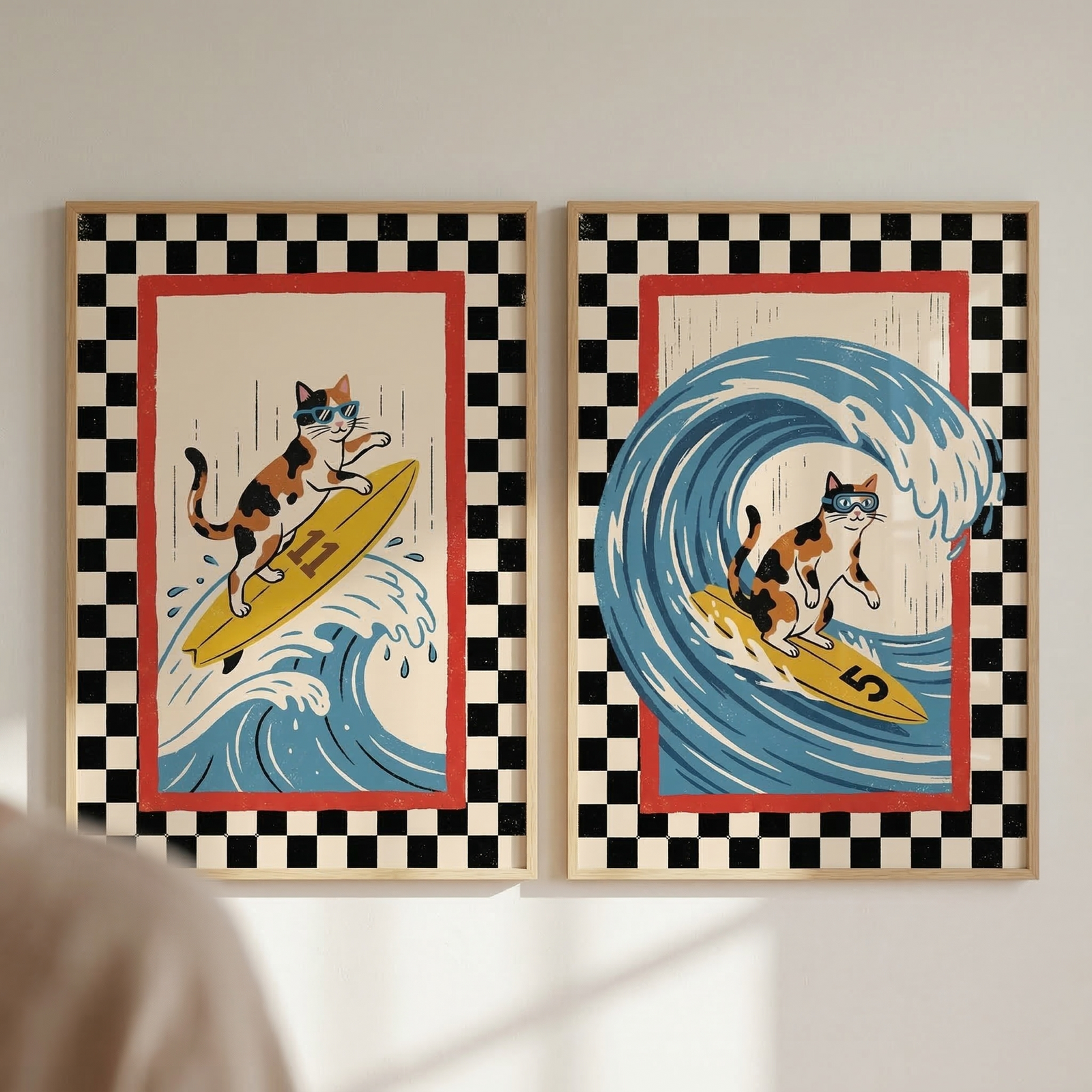 Two wood-framed illustrations hang side-by-side on a light-colored wall. Each artwork depicts a calico cat wearing blue goggles while surfing on a yellow board. Both pieces feature a wide black-and-white checkered border and a thin red interior frame. In the left frame, the cat rides a board numbered 11 down the face of a blue wave. In the right frame, the cat is shown inside the barrel of a large, curling blue wave on a board numbered 5.