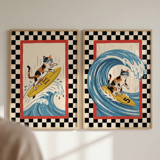Two wood-framed illustrations hang side-by-side on a light-colored wall. Each artwork depicts a calico cat wearing blue goggles while surfing on a yellow board. Both pieces feature a wide black-and-white checkered border and a thin red interior frame. In the left frame, the cat rides a board numbered 11 down the face of a blue wave. In the right frame, the cat is shown inside the barrel of a large, curling blue wave on a board numbered 5.