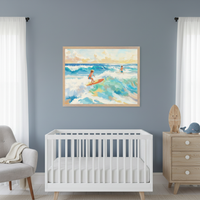 Modern nursery with a white crib and light gray armchair. A large framed painting depicts two children surfing on vibrant blue and green waves under a sunny sky, hanging above the crib. A light wood dresser with a surfboard and whale plush is on the right.