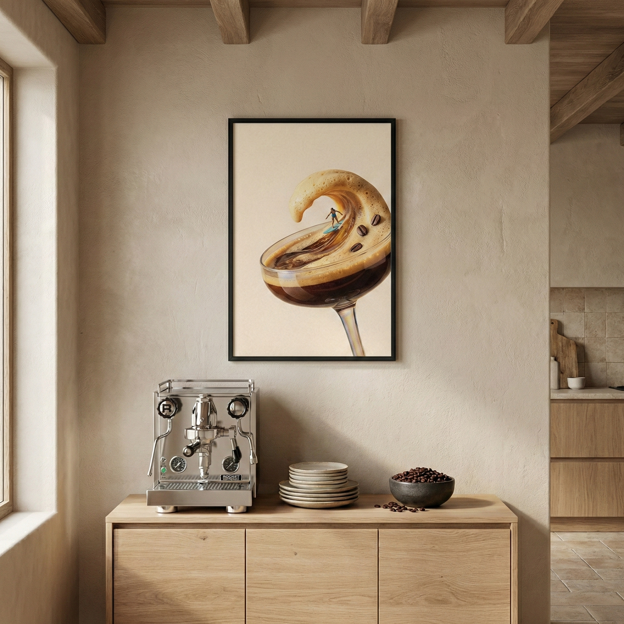 An eye-level indoor shot shows a light wood console table against a beige textured wall in a minimalist room. Resting on the table's surface are a silver, professional-style espresso machine, a stack of small ceramic plates, and a dark bowl overflowing with roasted coffee beans. Centered on the wall above the table is a framed vertical piece of art in a black frame. The artwork displays a surreal image: a glass of espresso where the light-colored foam forms a large, curling wave. 